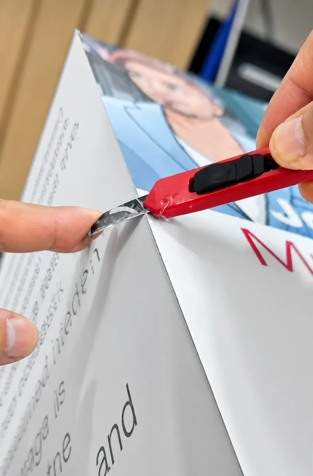 Hands trimming excess material from the folded edge of a printed display panel with a red utility knife