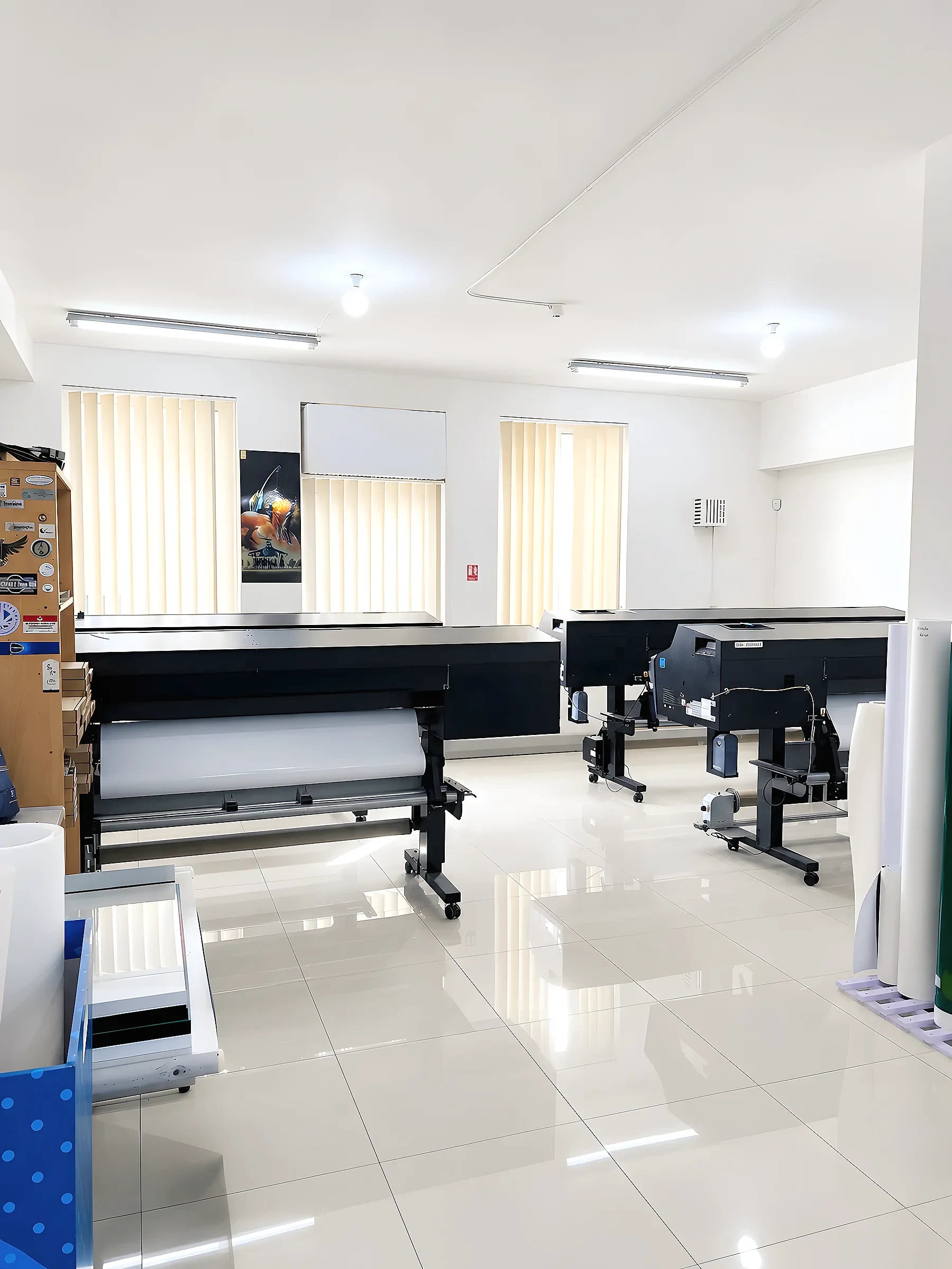 Two wide-format printers loaded with roll media in a bright tiled print room, with media rolls stacked alongside