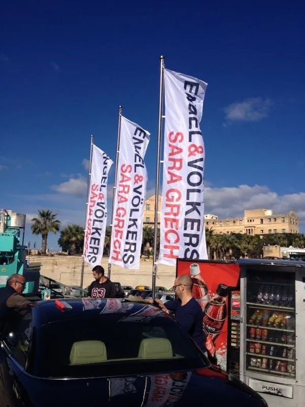 Row of white printed flags flying from poles along a seafront above parked vehicles