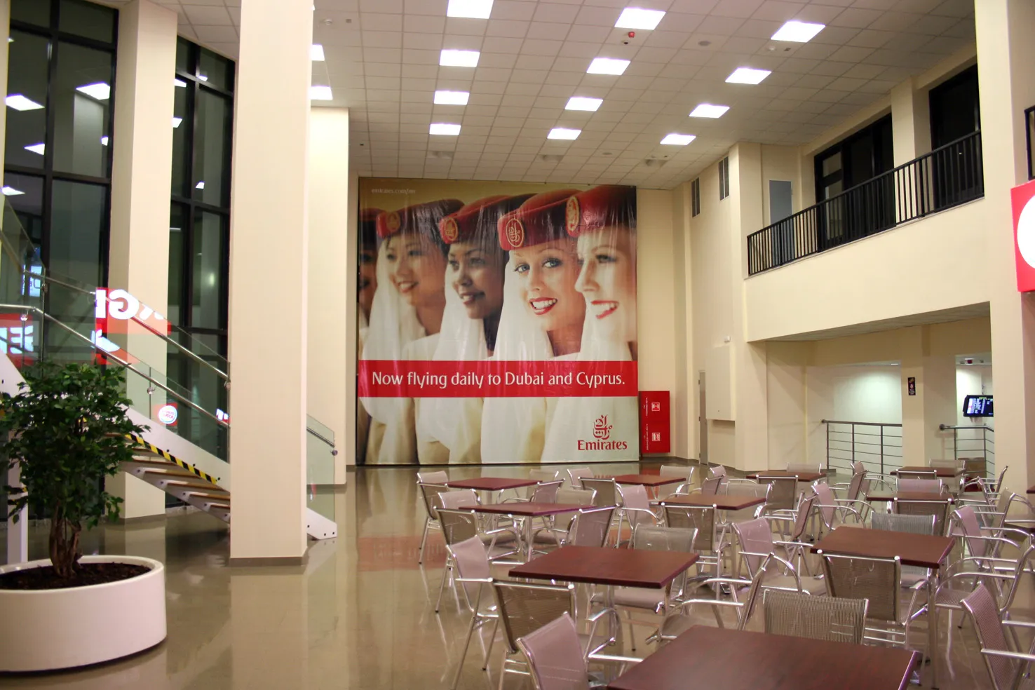 Large photographic wall graphic of a row of portraits high on the end wall of a double-height atrium, with a red message band and logo beneath