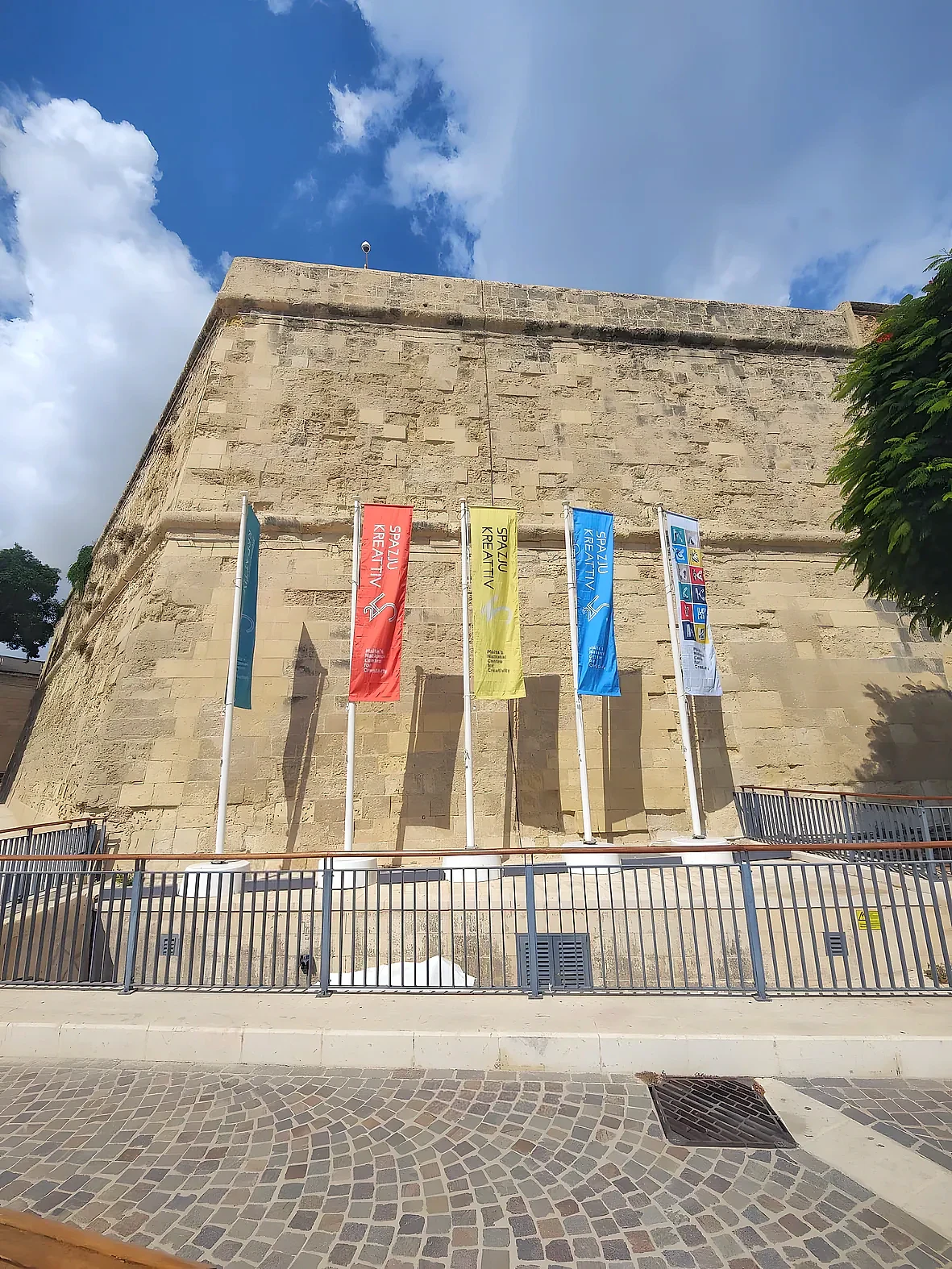 Five coloured banner flags on angled poles against a limestone bastion wall under a blue sky