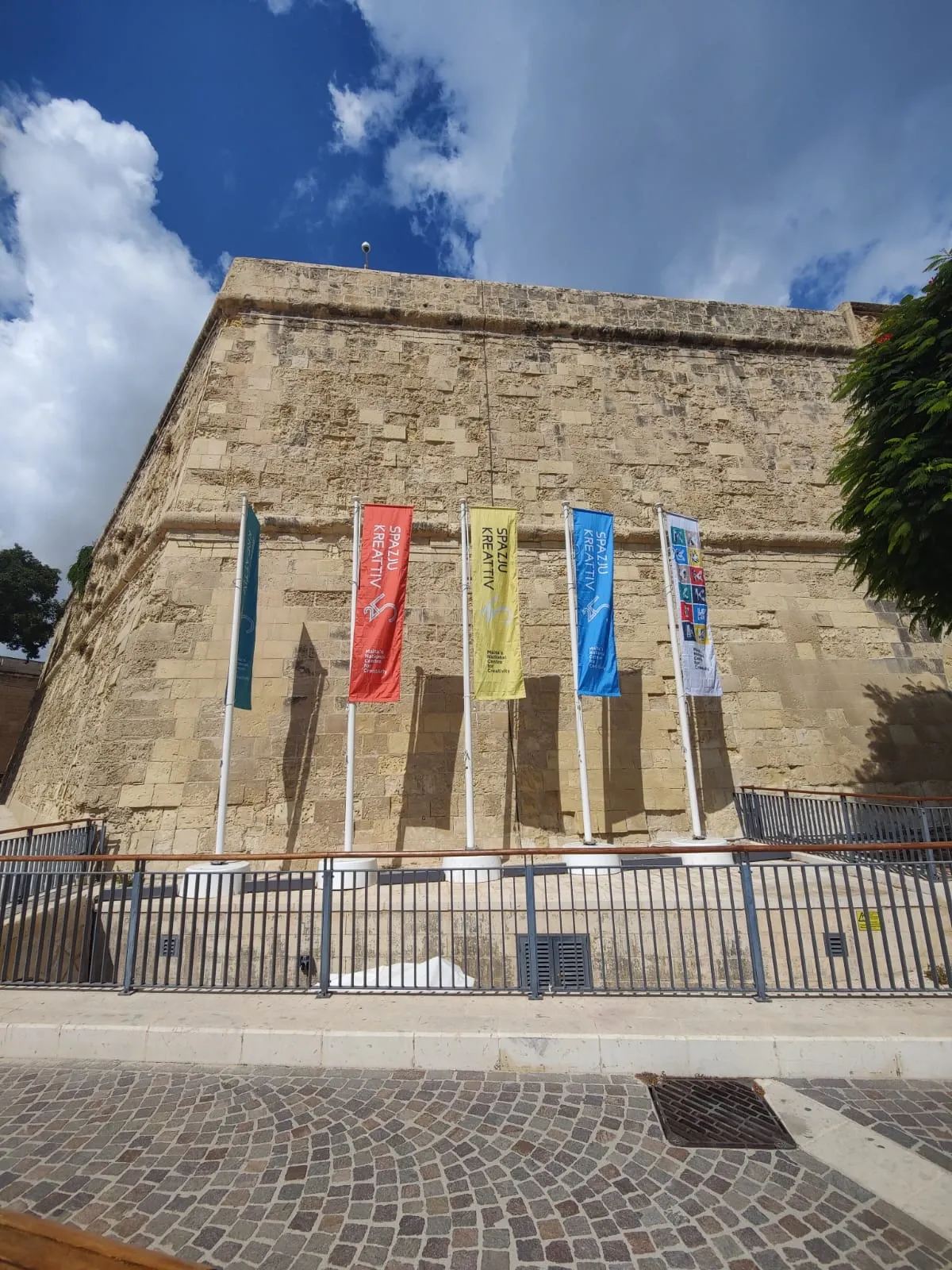 Five coloured banner flags on angled poles against a limestone bastion wall under a blue sky