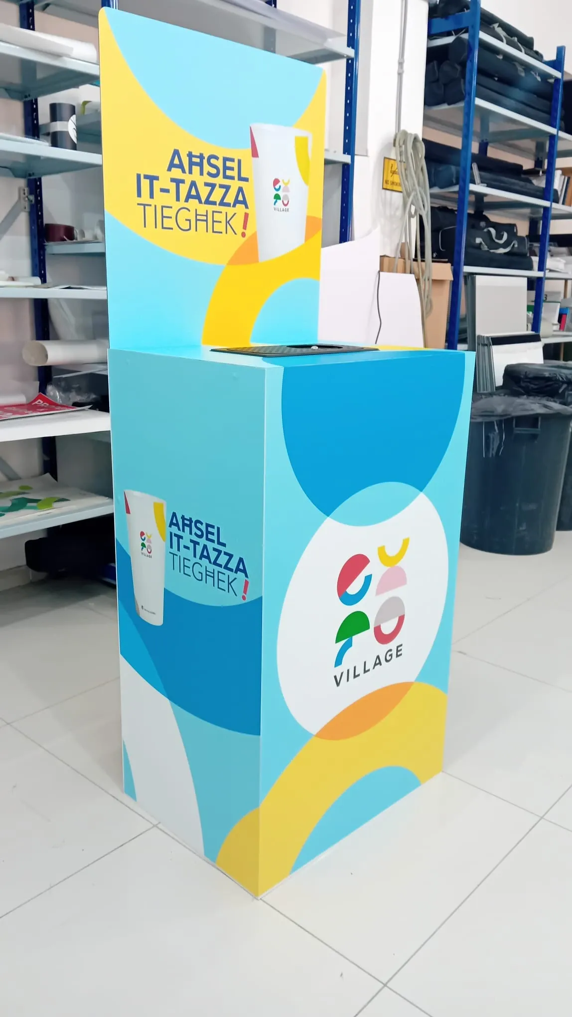 Blue and yellow branded cup collection bin with a raised printed header, in a storage area