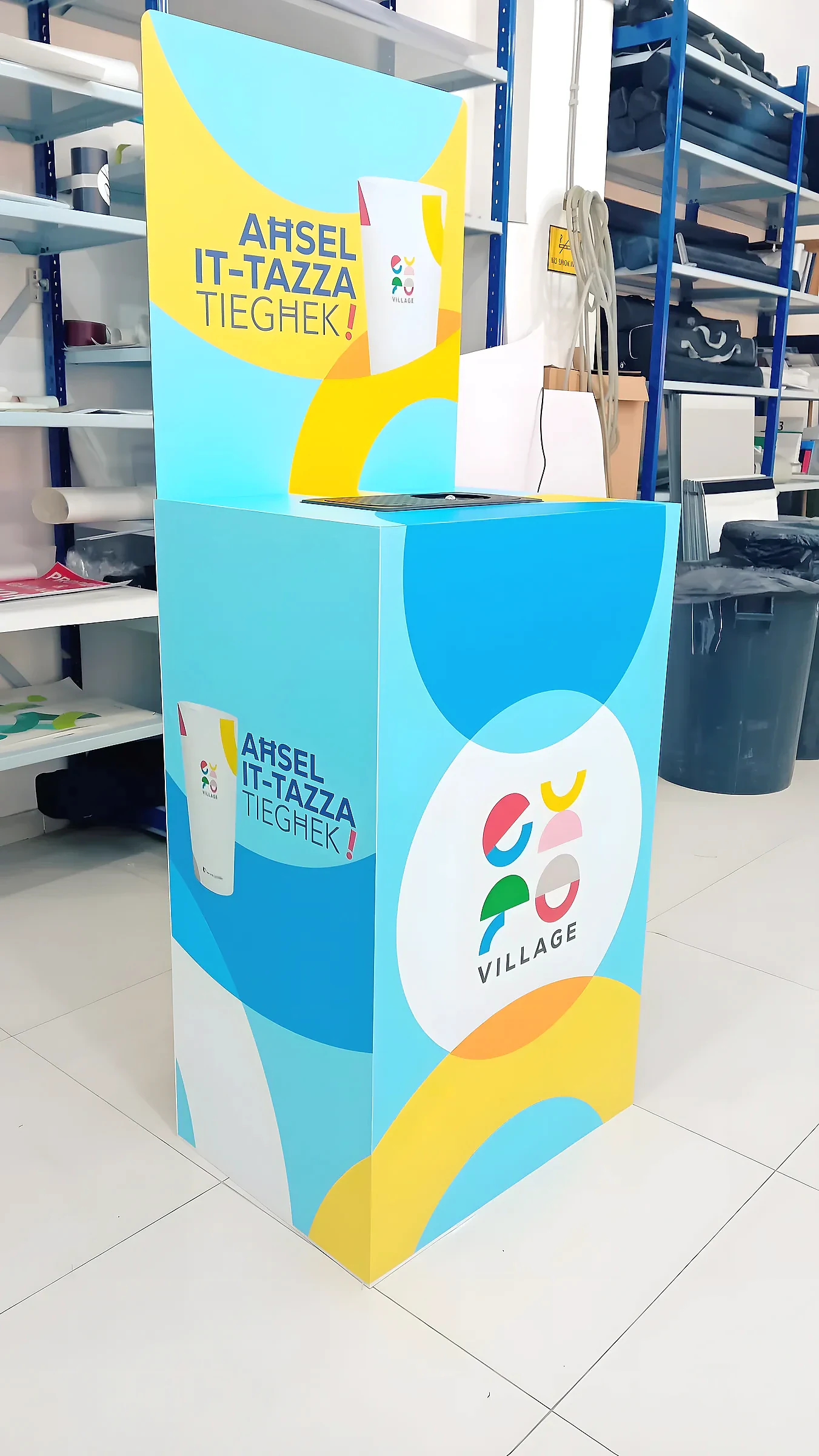 Blue and yellow branded cup collection bin with a raised printed header, in a storage area