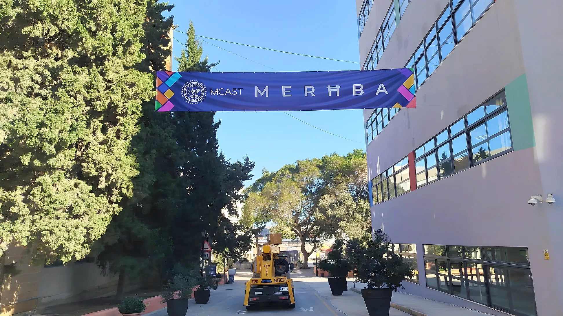 Purple welcome banner strung across a campus road between trees and a multi-storey building