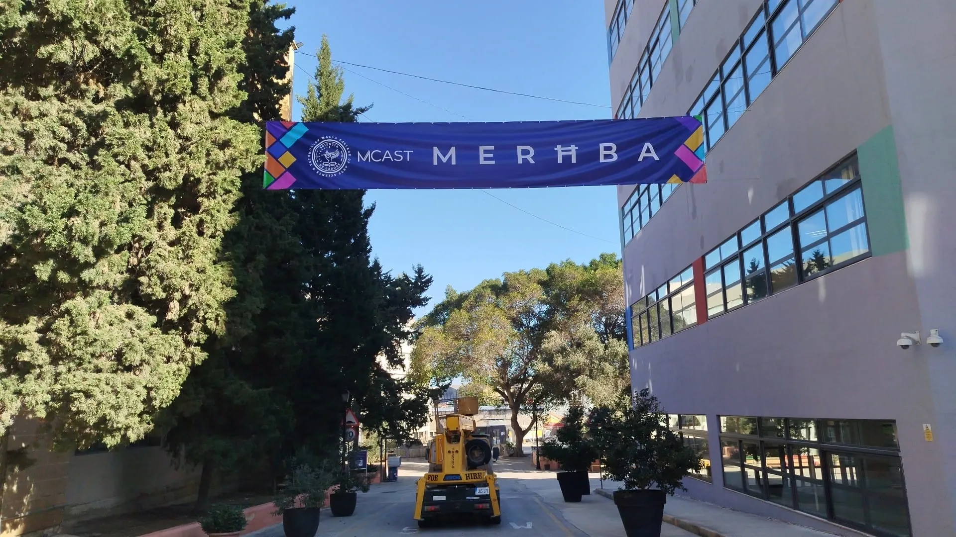 Purple welcome banner strung across a campus road between trees and a multi-storey building