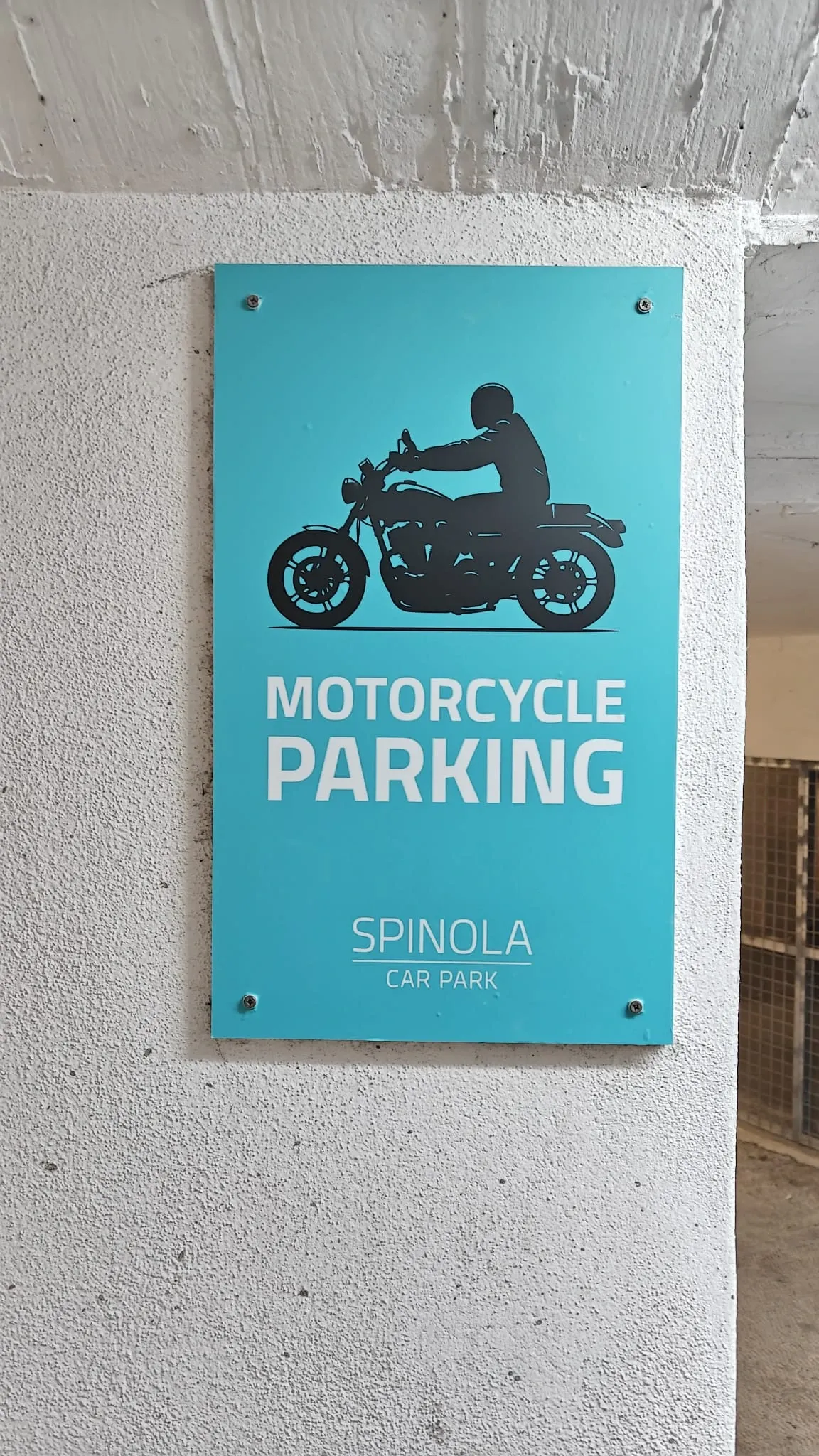 Turquoise motorcycle parking sign screw-fixed to a white concrete car park wall