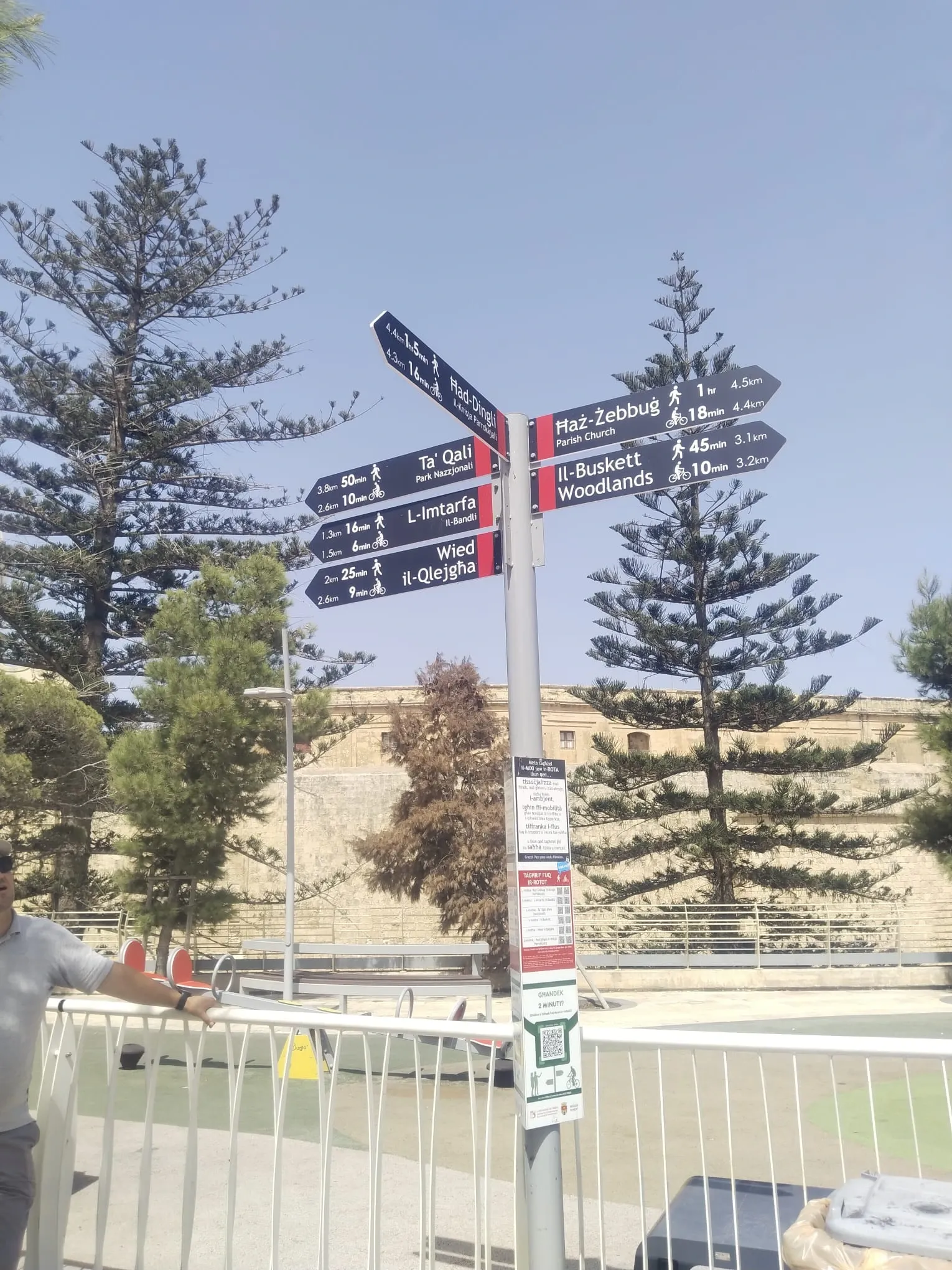 Fingerpost with eight destination arms on a tall post against pine trees and a bastion wall