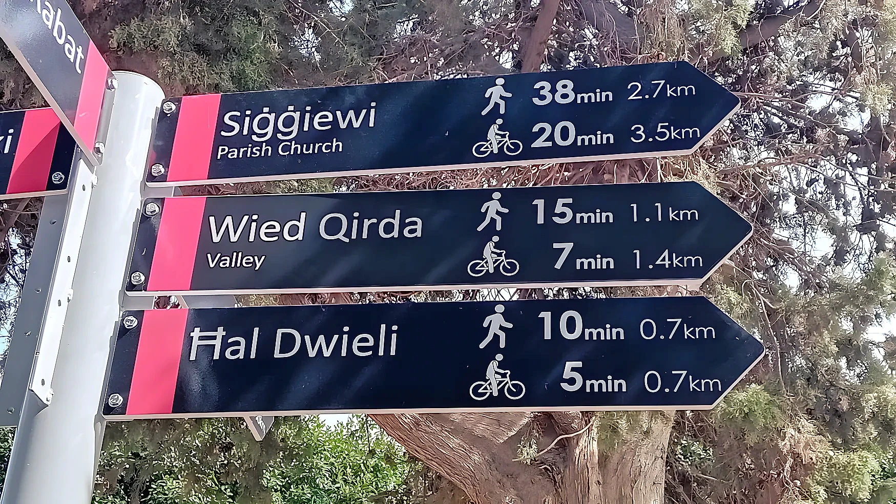 Three black fingerpost arms giving walking and cycling times to Maltese villages and a valley