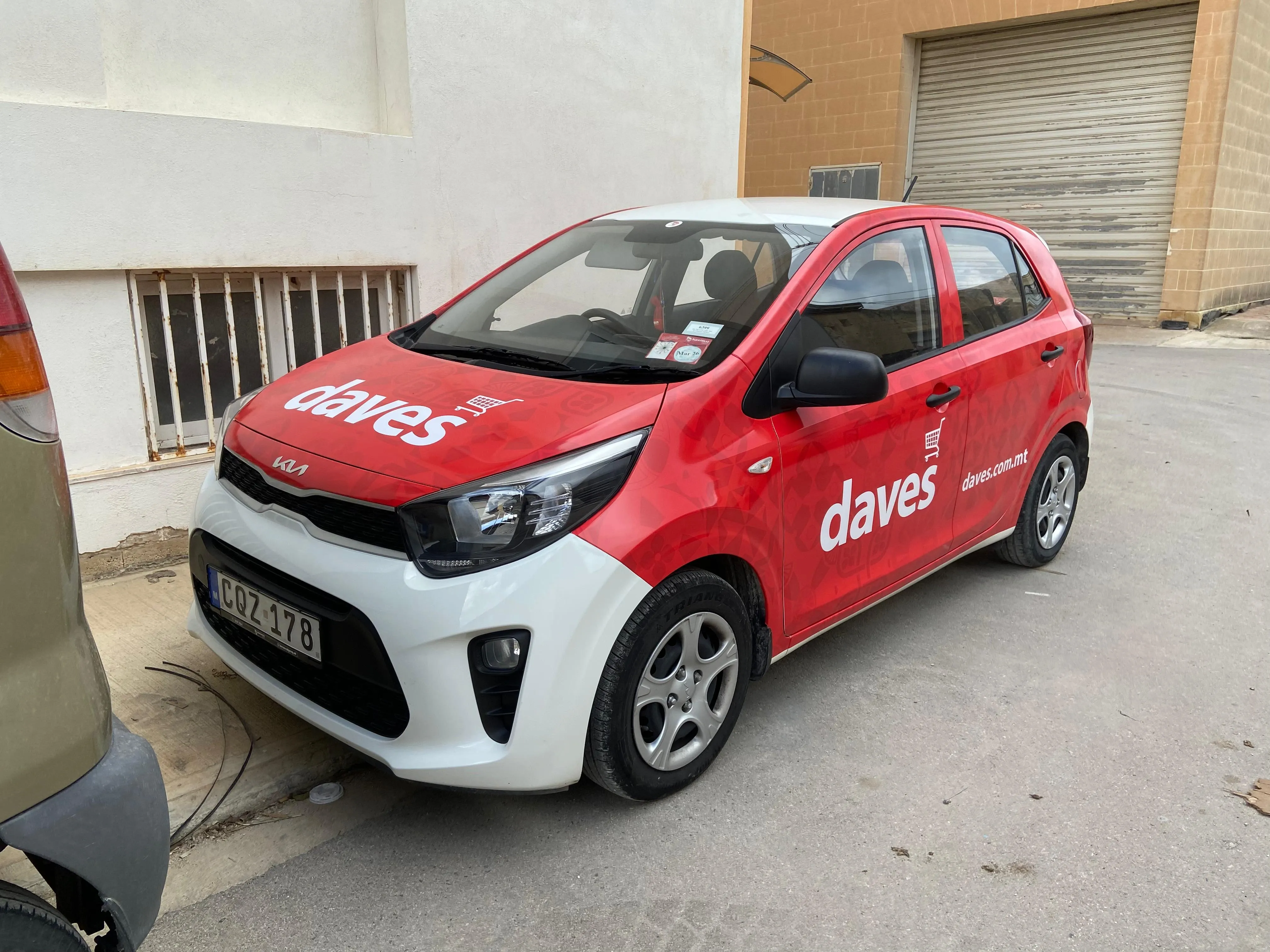 Red and white wrapped city car with a brand name on the bonnet and door, parked by a warehouse