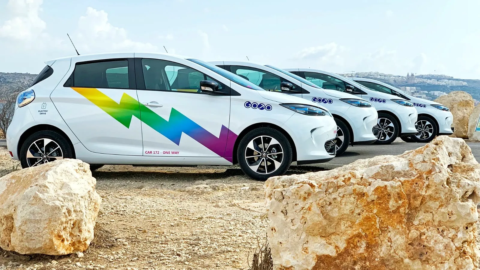 Row of white electric cars with matching multicolour geometric graphics beside a rocky shoreline