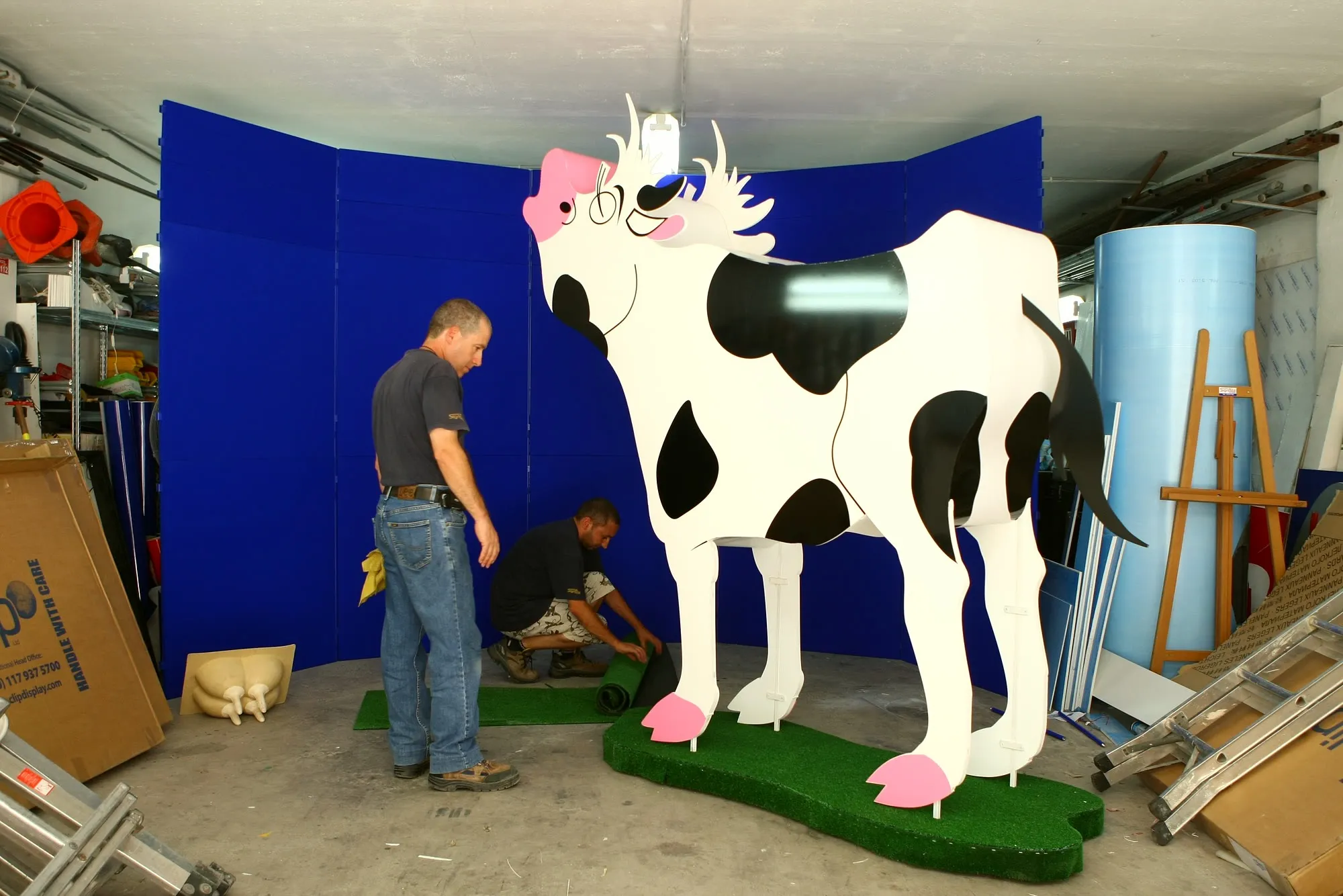 Workshop technician fitting the base of a full-size fabricated display figure against a blue screen