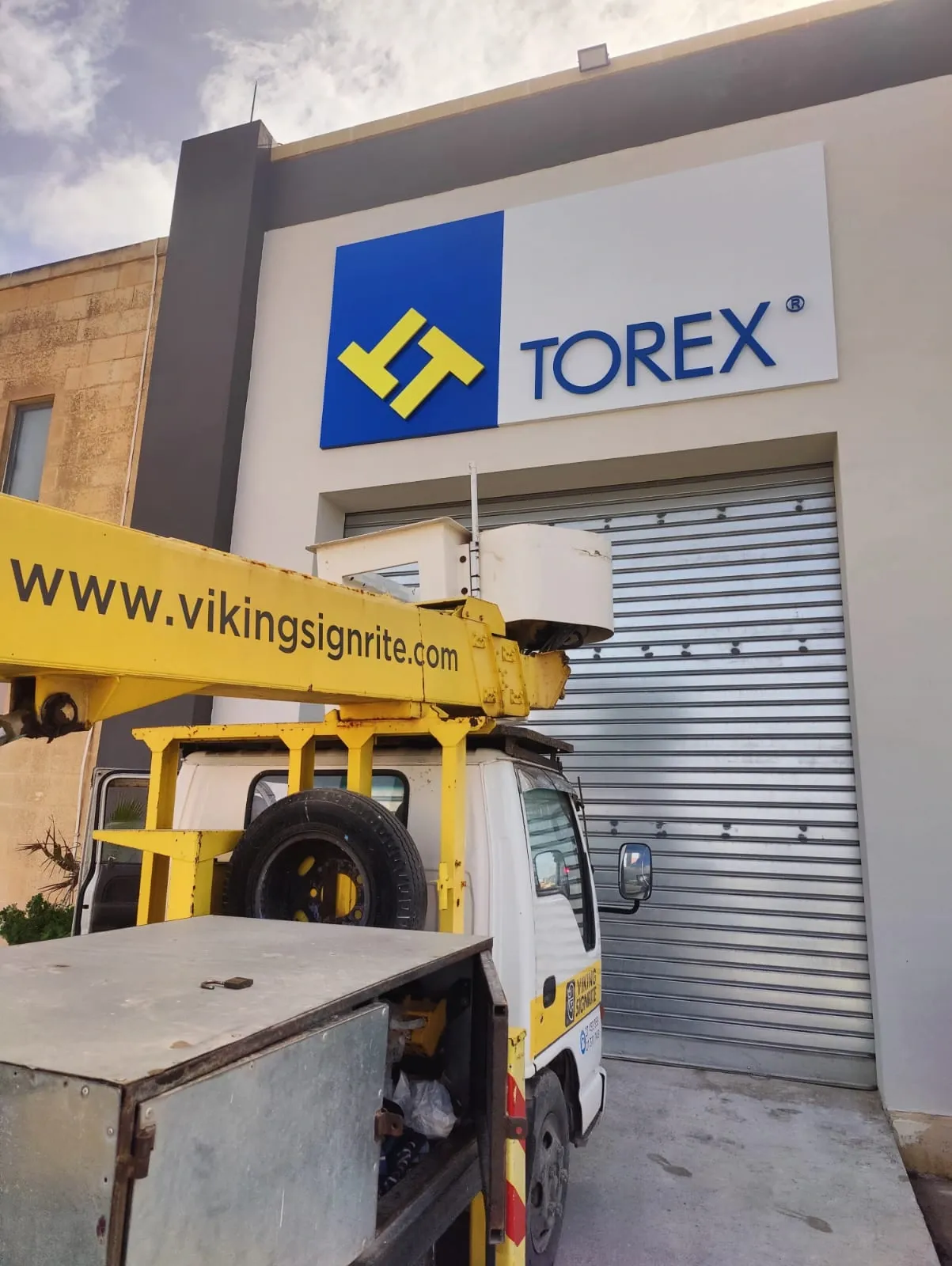 Blue and yellow logo panel with built-up lettering high on an industrial facade, with a cherry picker below