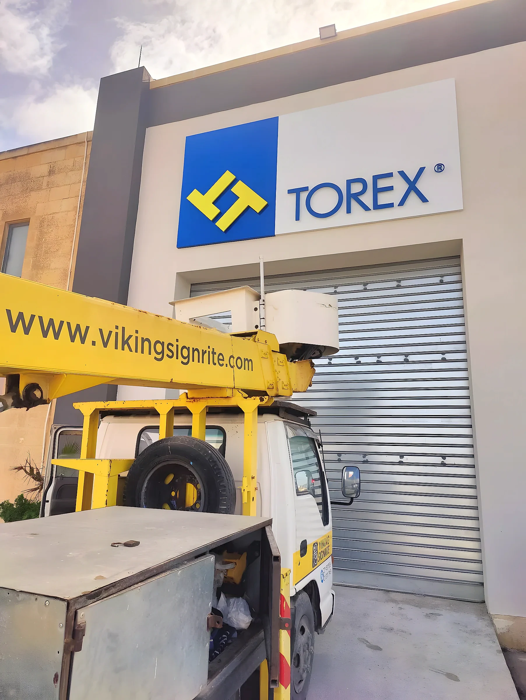 Blue and yellow logo panel with built-up lettering high on an industrial facade, with a cherry picker below