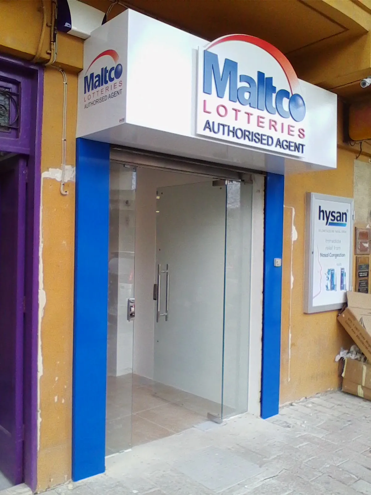 A white fascia box sign above a glazed shop door with a smaller matching box projecting from the corner, on a yellow rendered building