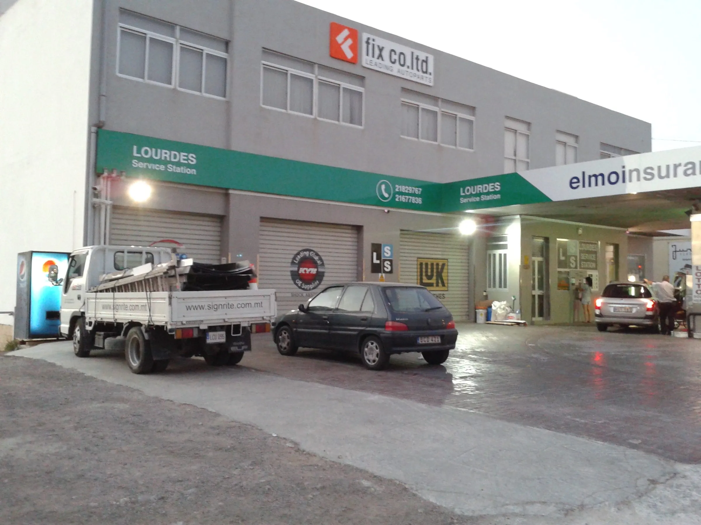 Single-storey forecourt retail building with a green and white fascia sign and cars parked in front