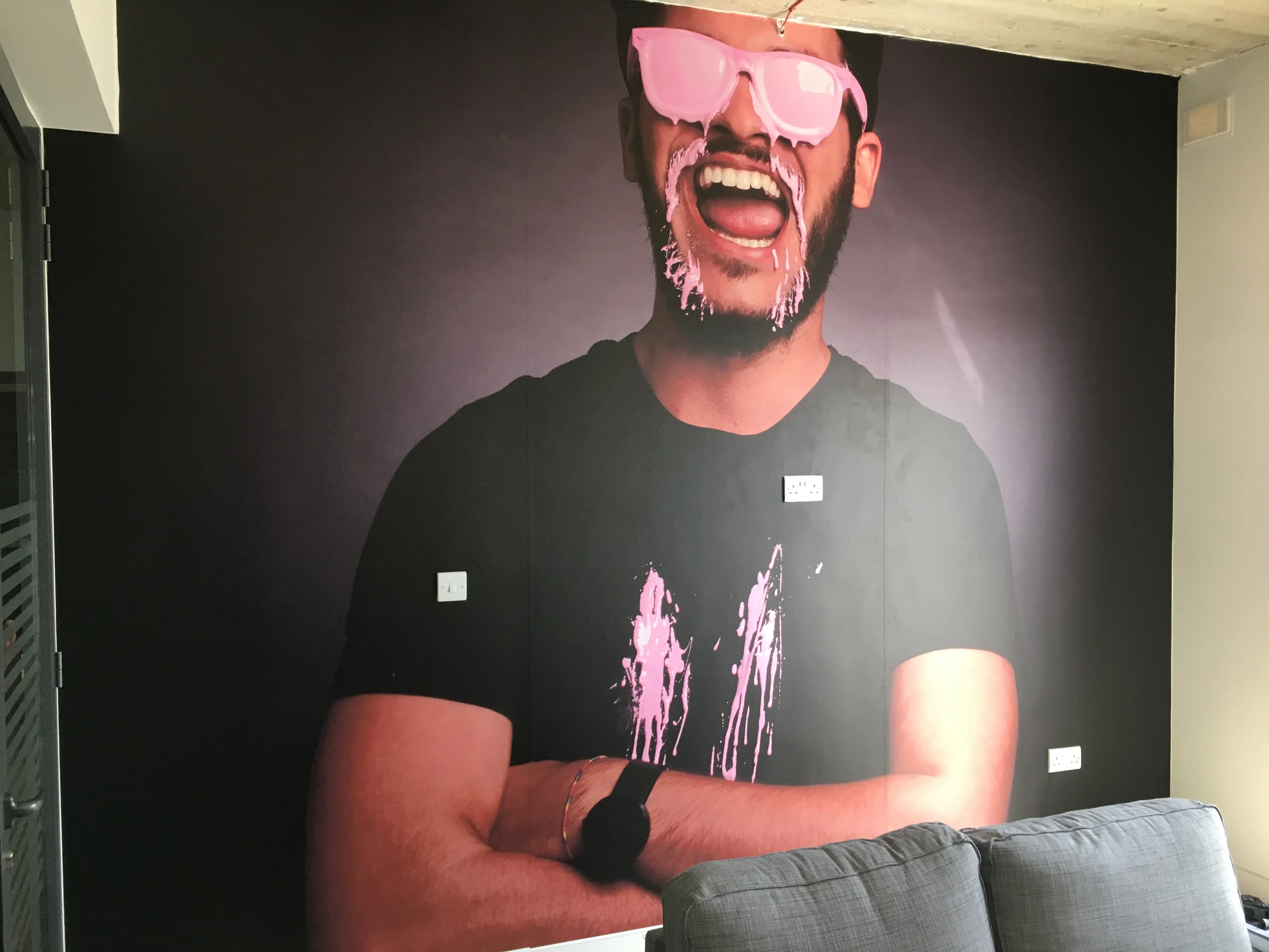 Very large photographic portrait of a laughing figure filling an interior wall floor to ceiling on a dark ground, with sockets and a switch cut around