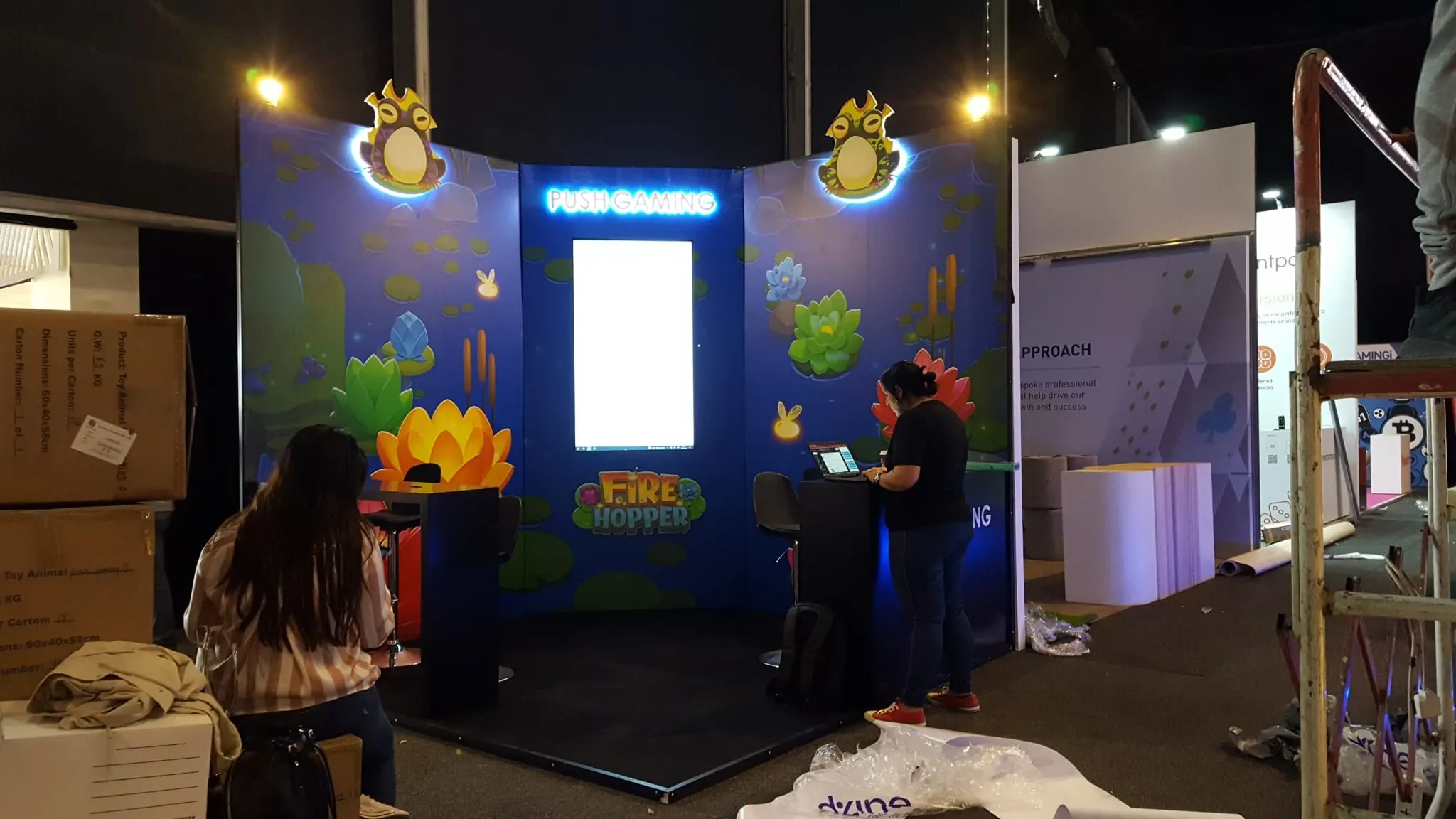 Illustrated gaming exhibition stand with lit character panels and a central lightbox being set up on a show floor