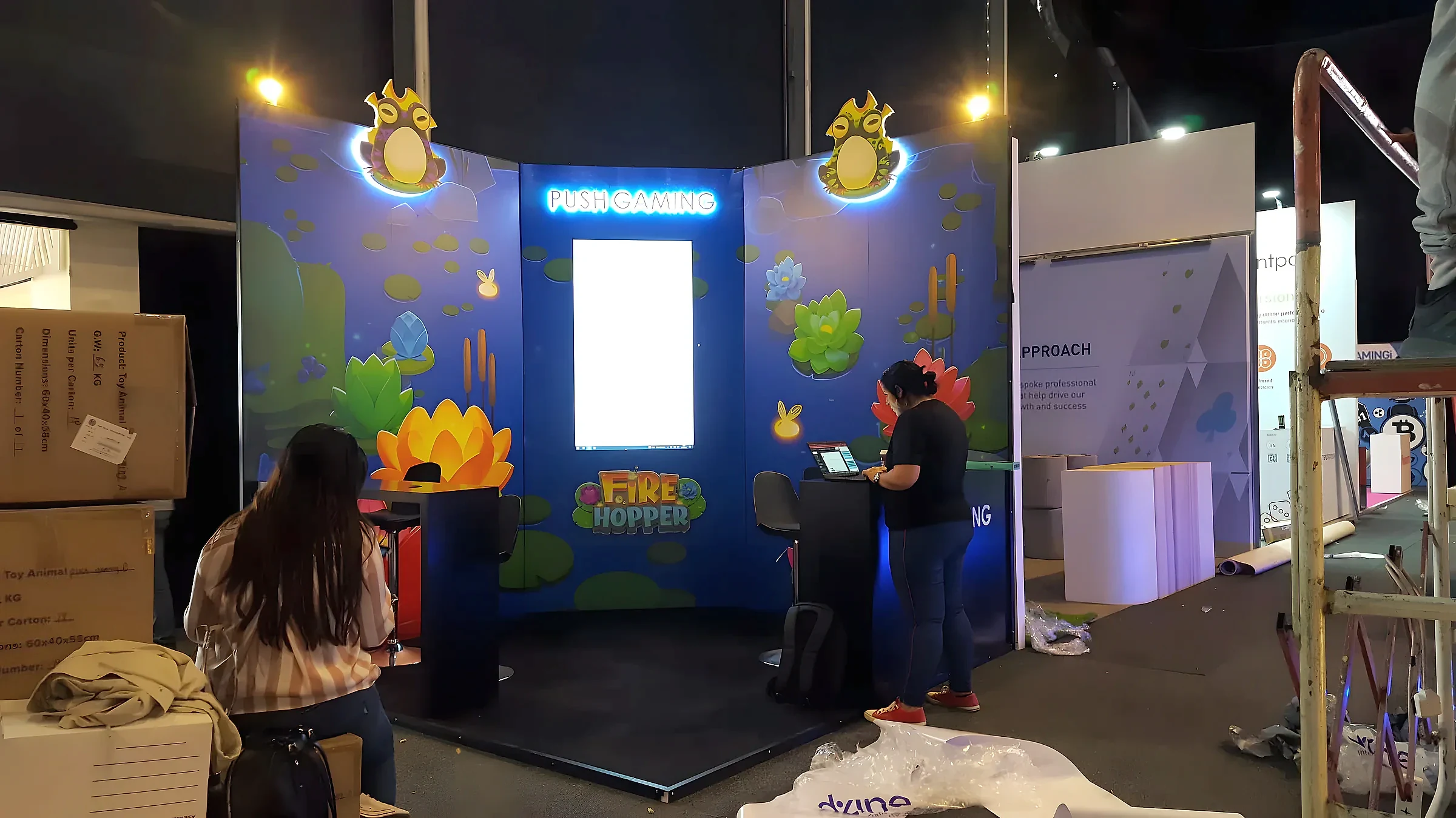 Illustrated gaming exhibition stand with lit character panels and a central lightbox being set up on a show floor