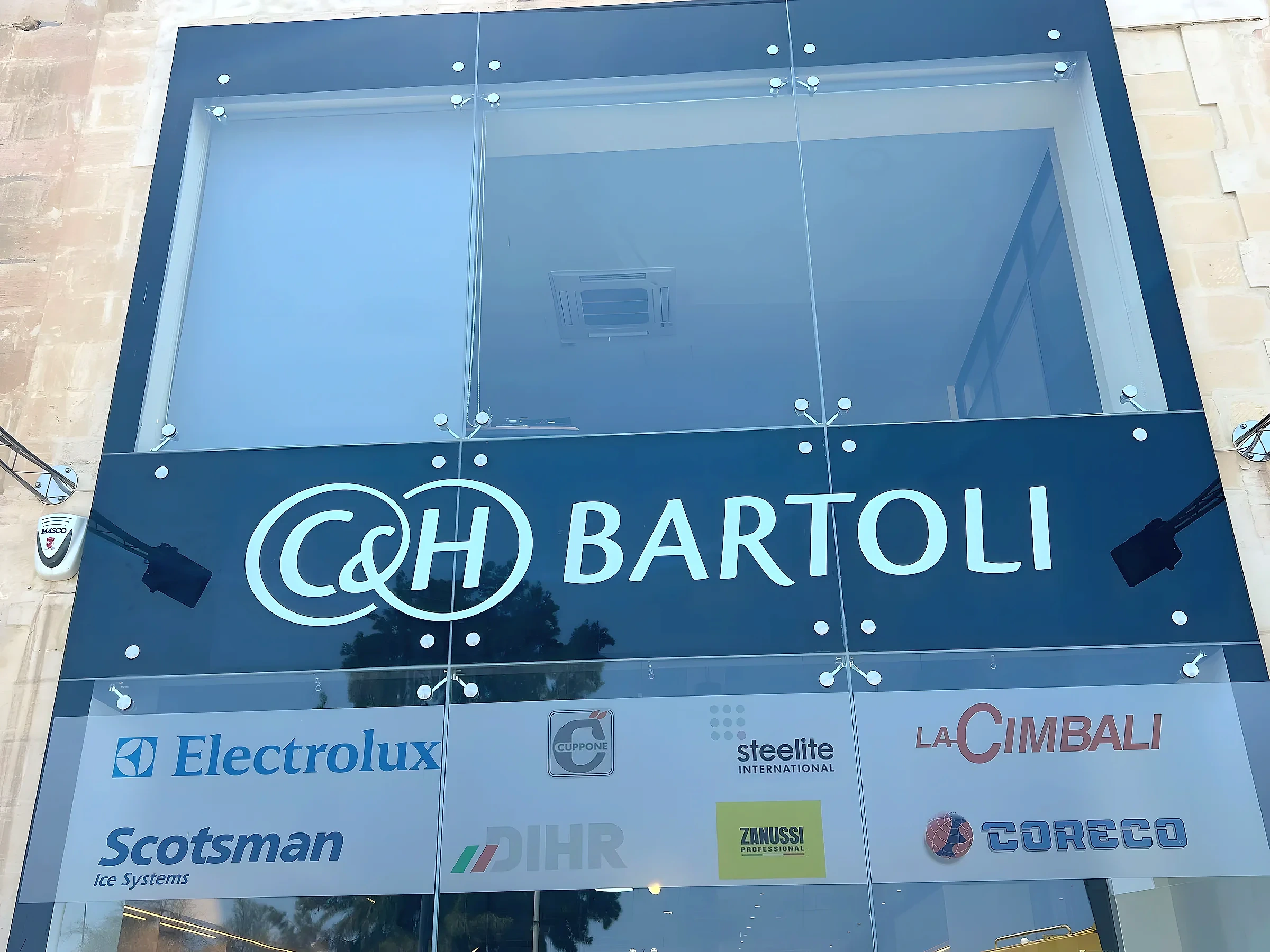 Dark fascia sign with large built-up lettering above a row of supplier plates on a glazed building frontage