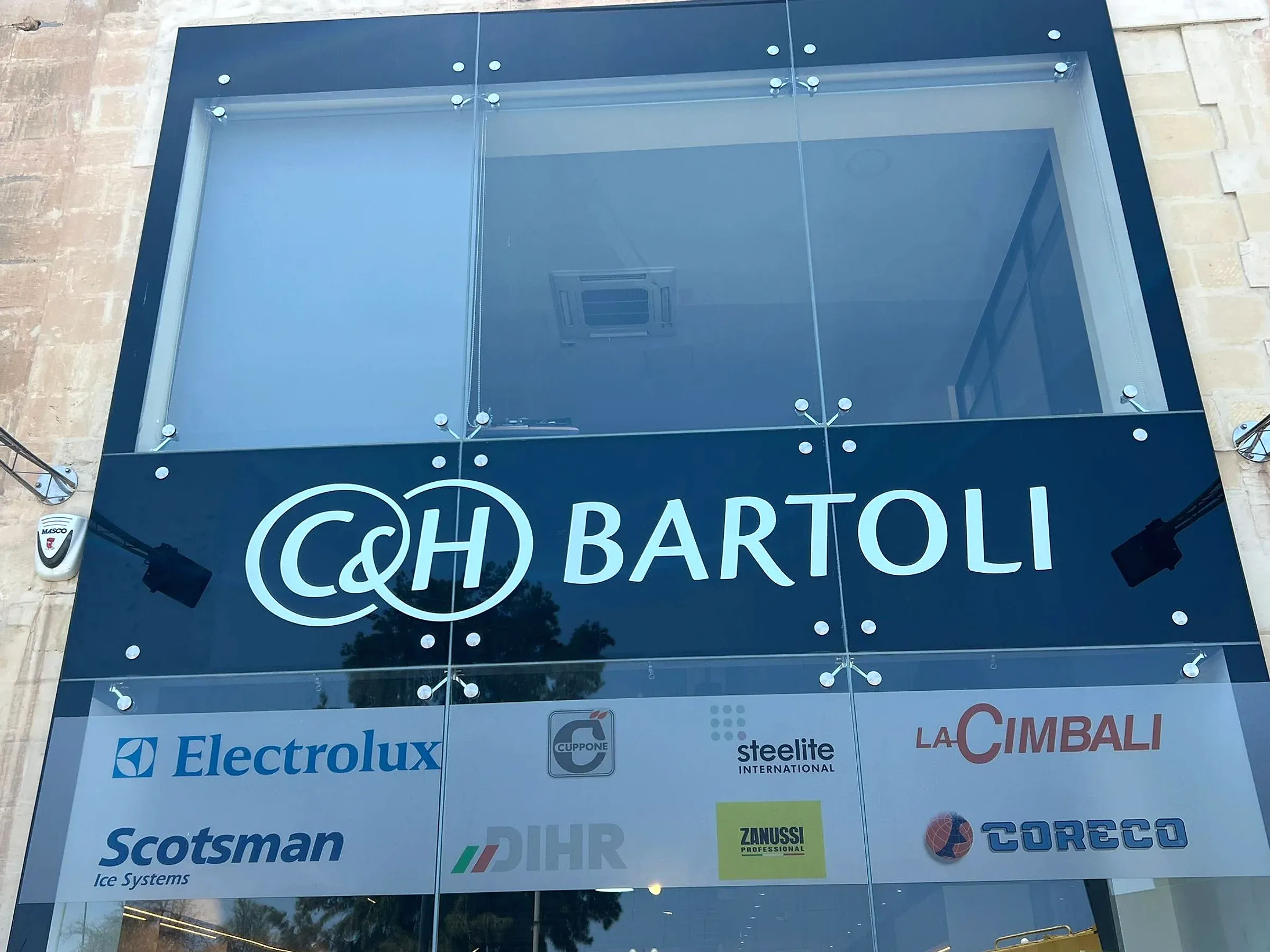 Dark fascia sign with large built-up lettering above a row of supplier plates on a glazed building frontage