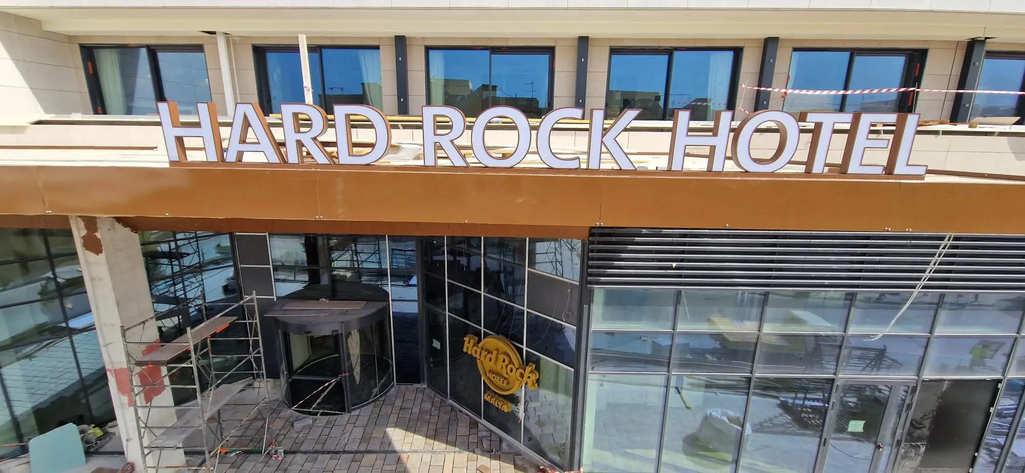 Large dimensional letters spelling a hotel name along the top of a fascia band on a multi-storey building, above a timber-clad entrance canopy
