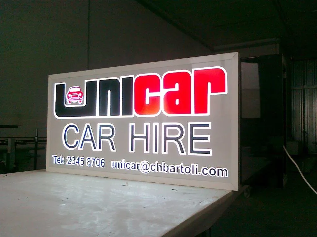 Large internally lit lightbox sign standing on castors in a dark workshop, its face glowing with coloured lettering and contact details