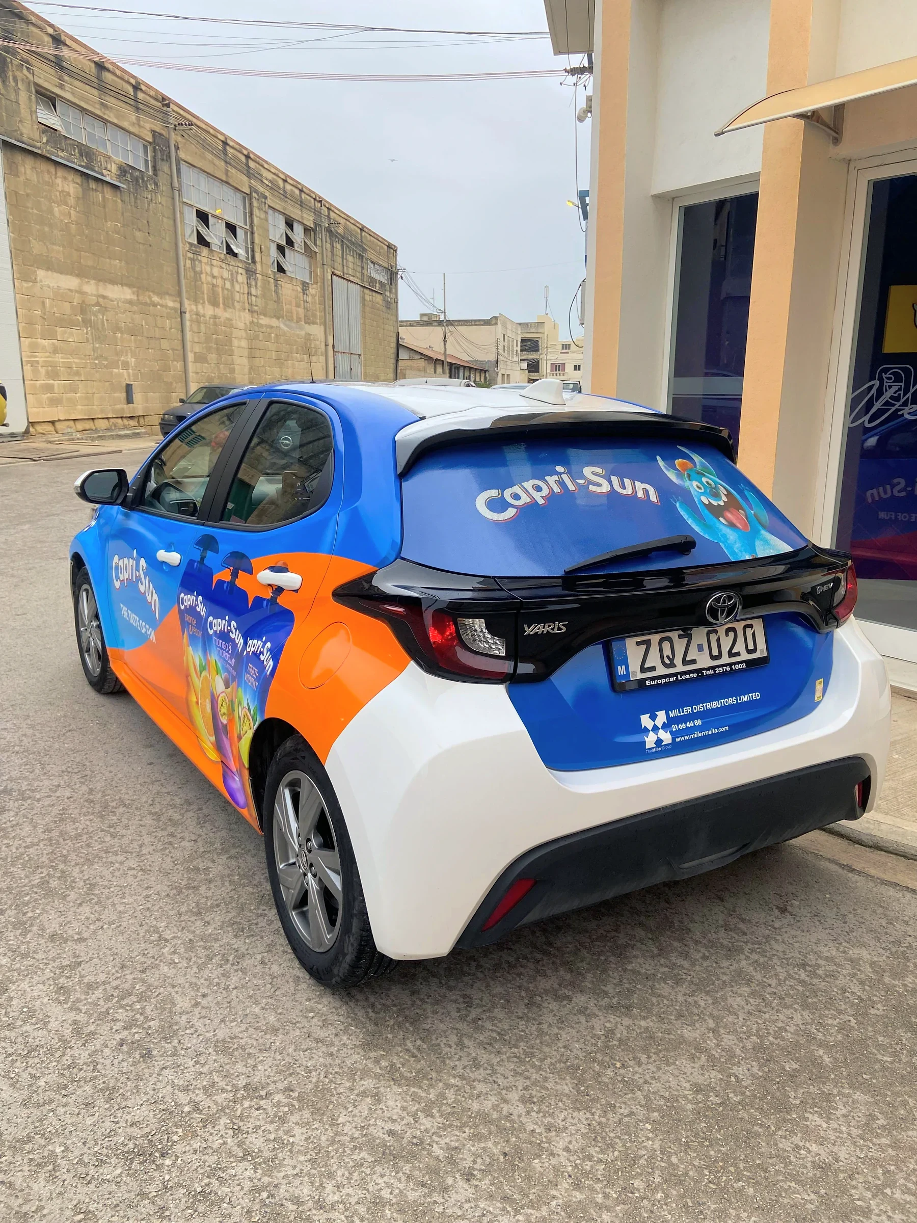 Blue and orange wrapped hatchback with a printed rear screen, parked outside a workshop