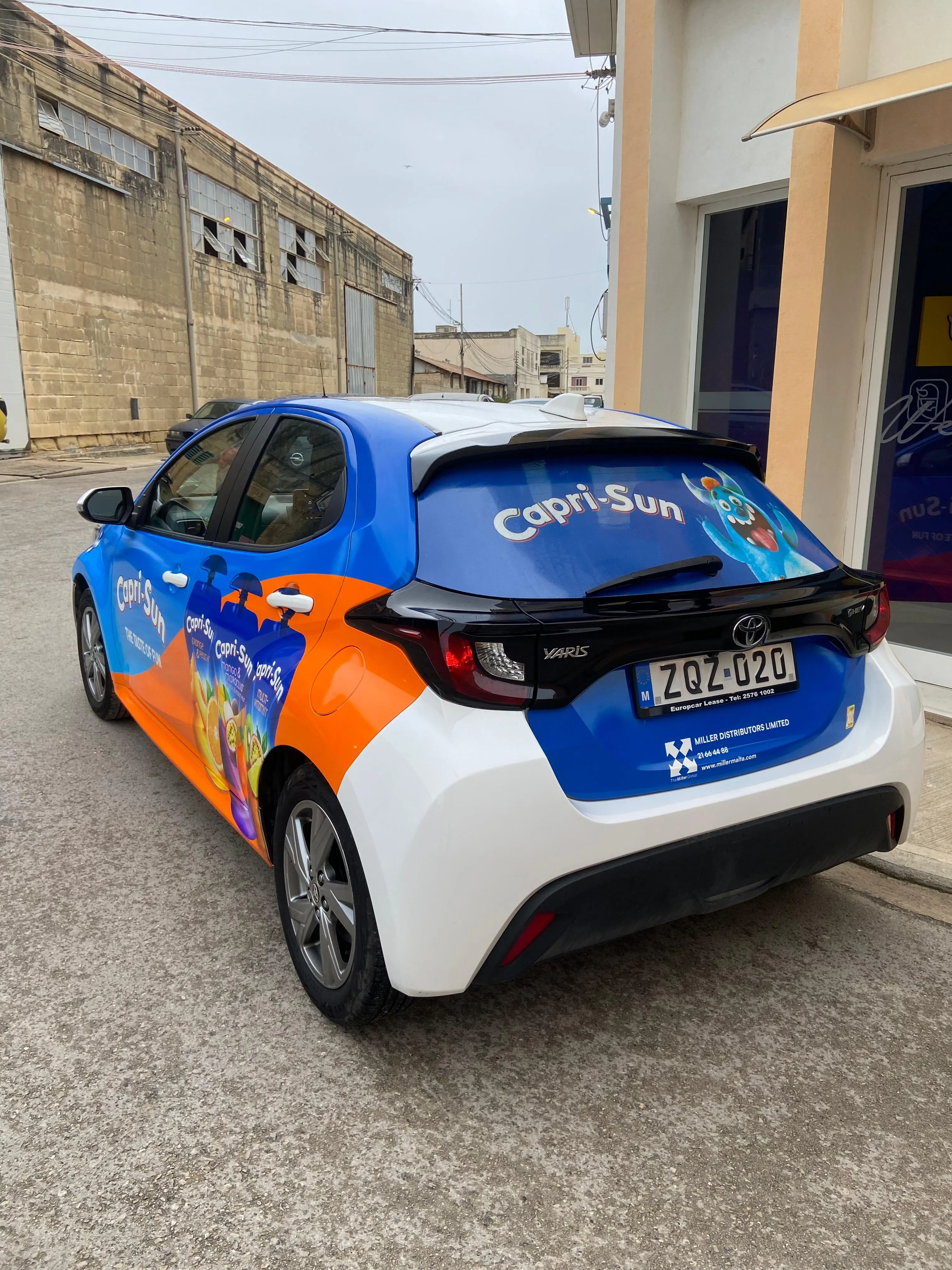 Blue and orange wrapped hatchback with a printed rear screen, parked outside a workshop