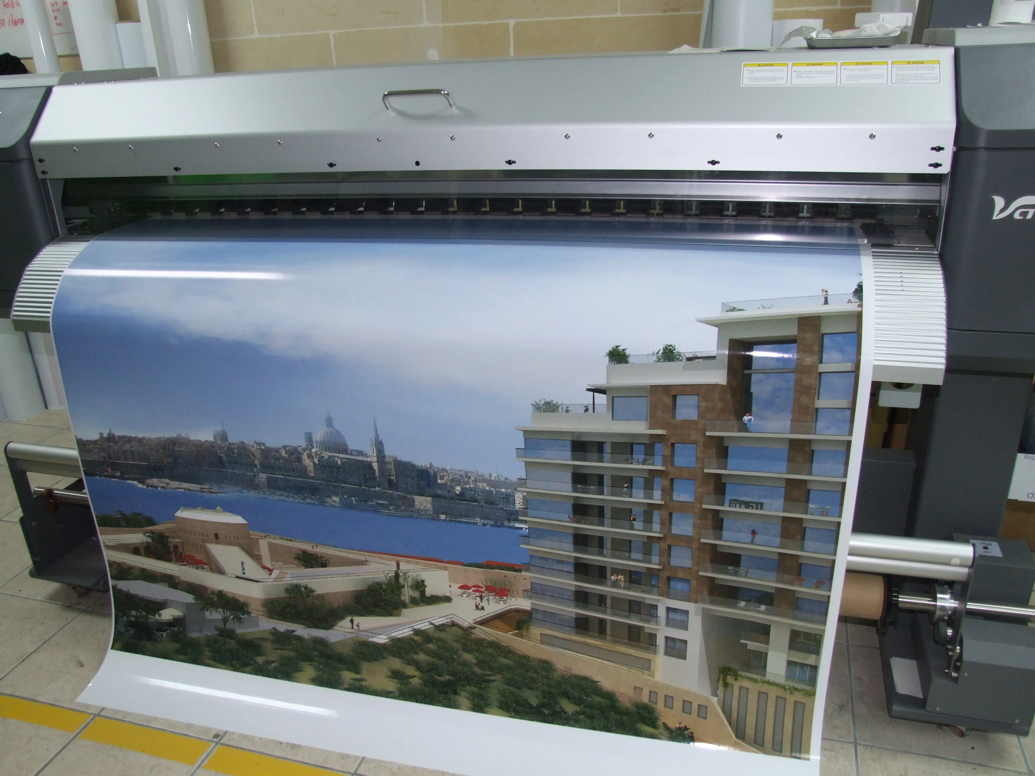 Wide-format printer producing a full-width architectural visualisation print, curving over the take-up roller in a workshop