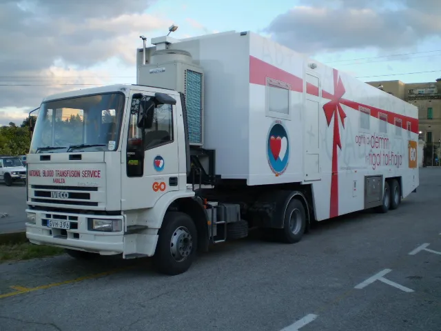Large mobile unit on a truck chassis, its white body carrying a sweeping ribbon graphic, an emblem, Maltese-language copy and lettering across the cab doors