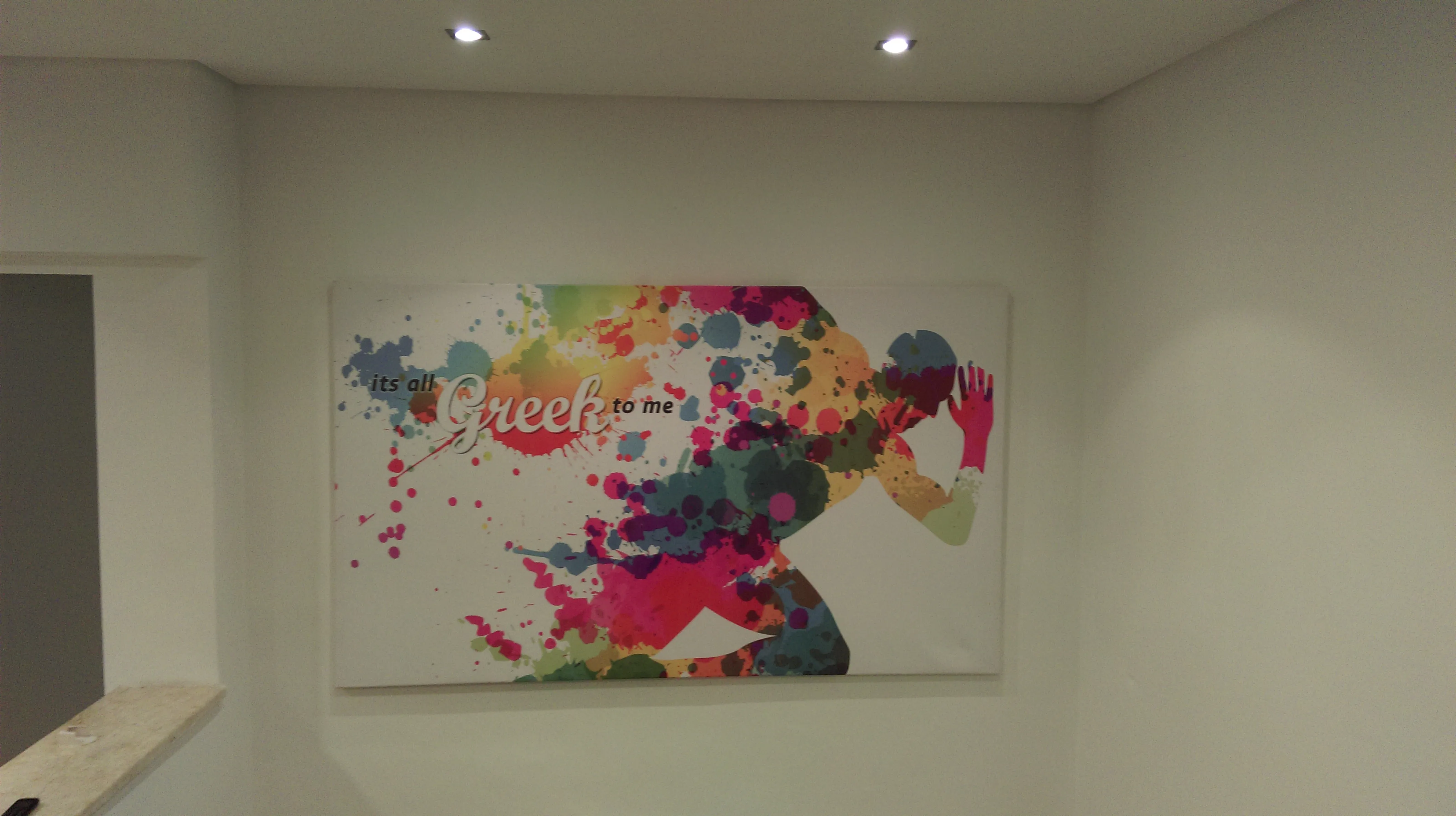 Large printed canvas panel mounted on an interior wall, showing a paint-splatter figure with script lettering, lit by two downlights