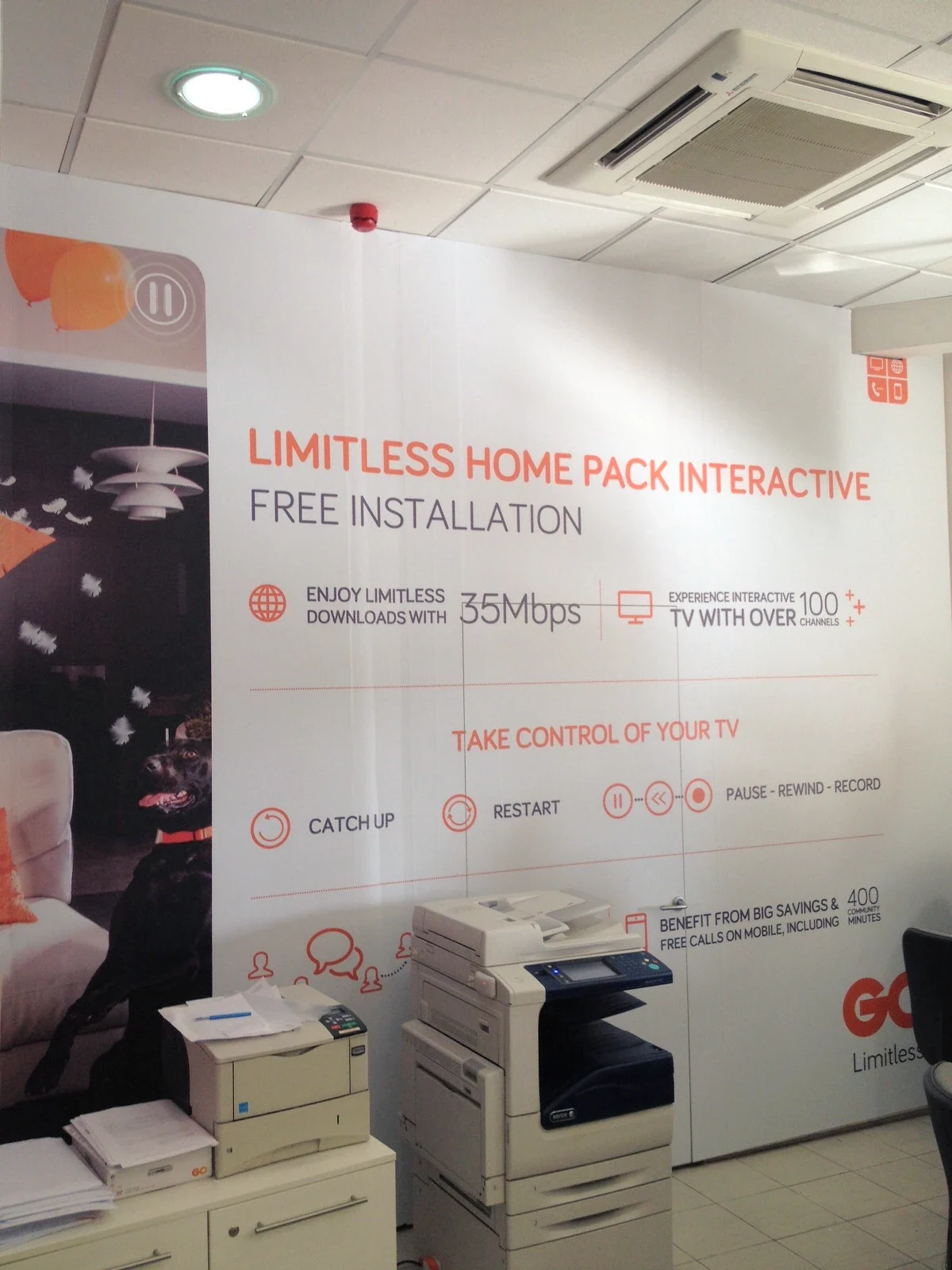 Floor-to-ceiling printed wall graphic in an office advertising a home internet and TV package, beside printers and paper trays