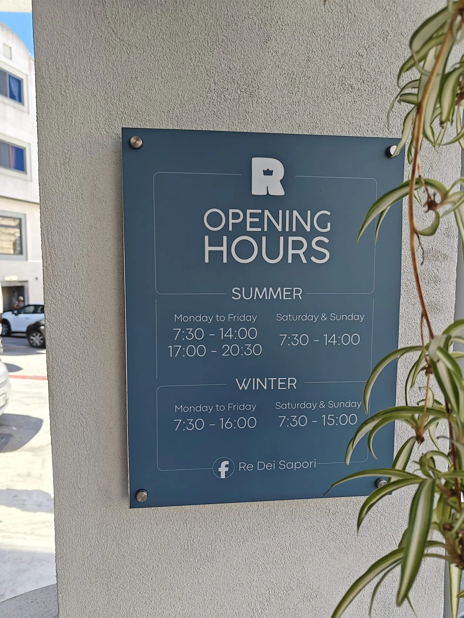 Blue-grey acrylic opening hours plaque on standoffs fixed to a rendered wall beside a plant