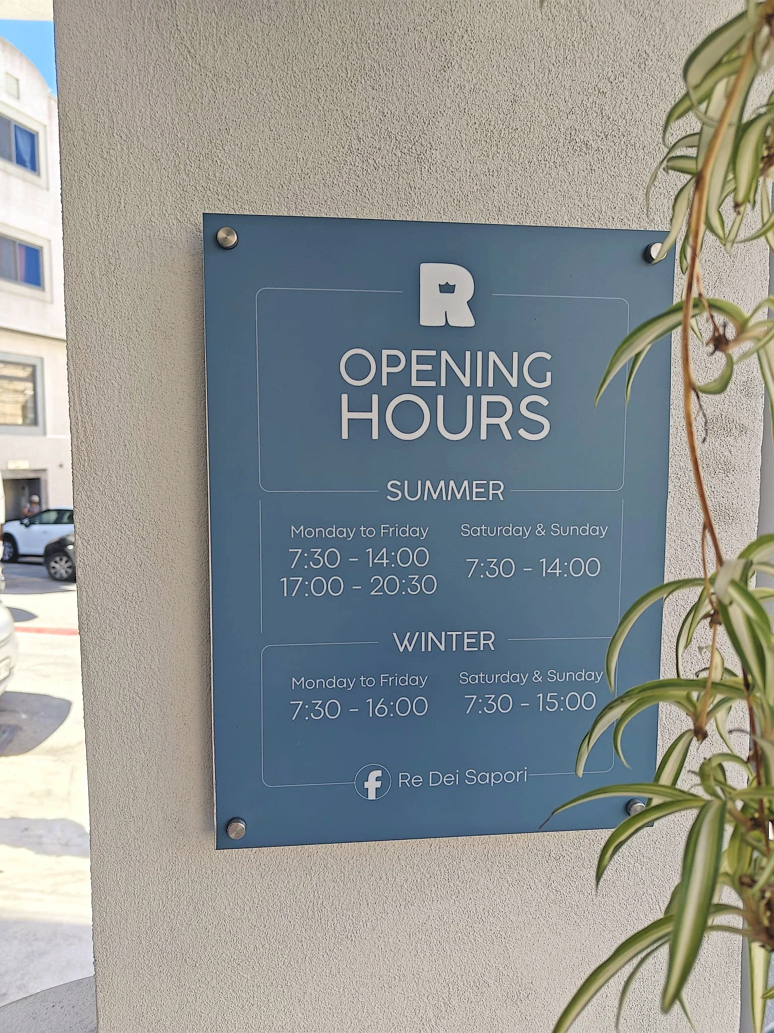 Blue-grey acrylic opening hours plaque on standoffs fixed to a rendered wall beside a plant