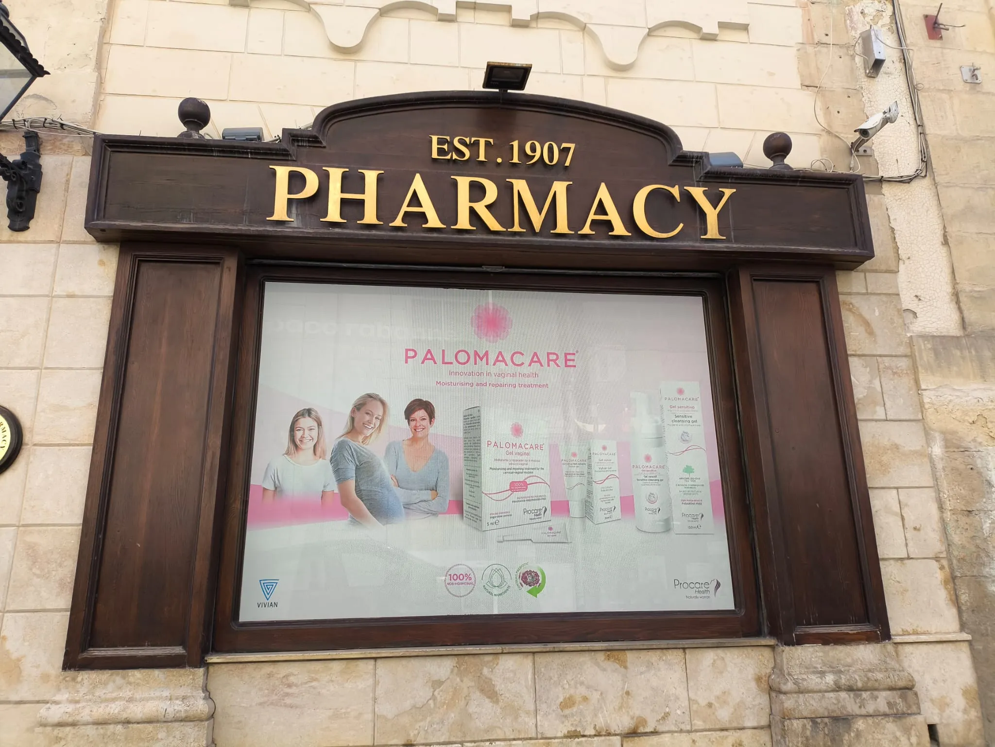 Printed product campaign panel fitted inside a traditional dark timber shopfront window beneath a gilded pharmacy fascia