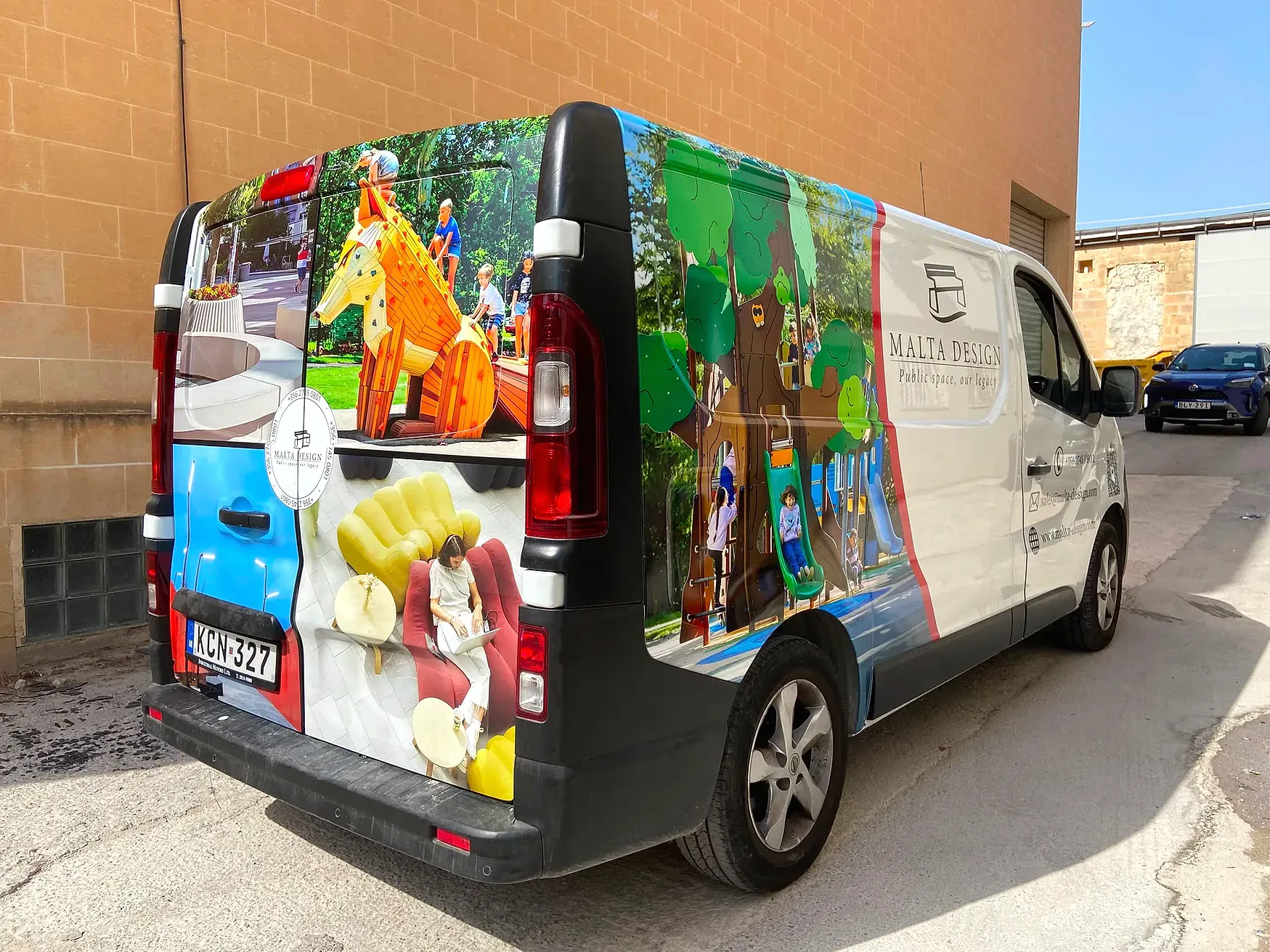 White panel van wrapped in photographs of outdoor play equipment and interior furniture, parked beside a stone wall