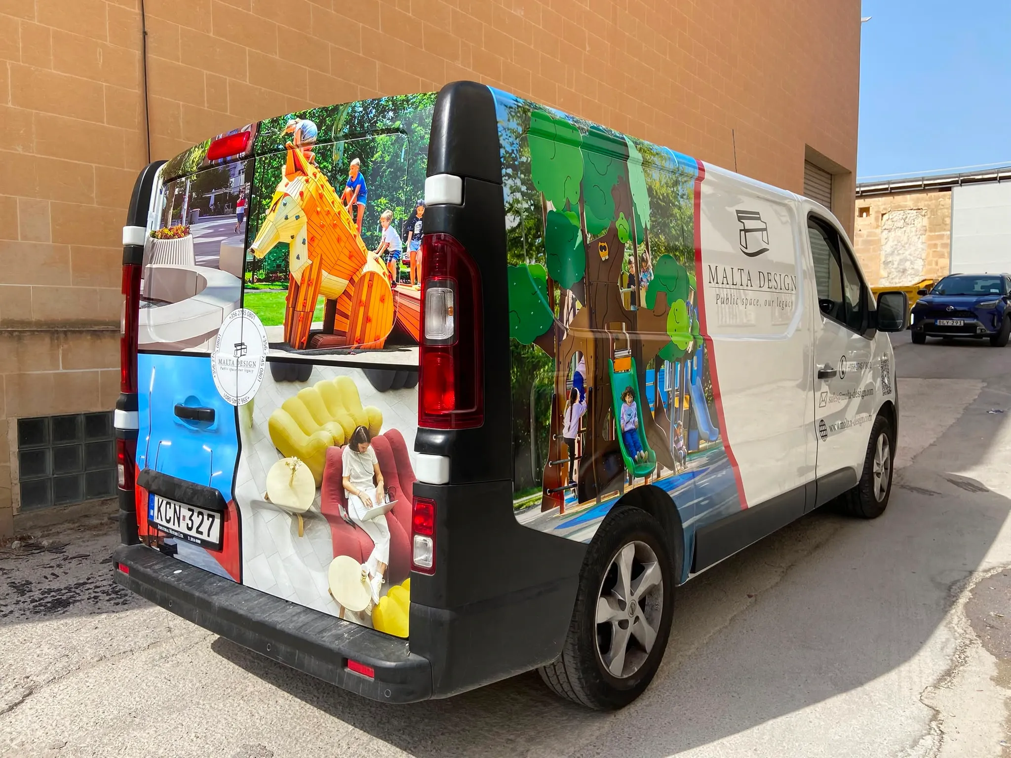 White panel van wrapped in photographs of outdoor play equipment and interior furniture, parked beside a stone wall