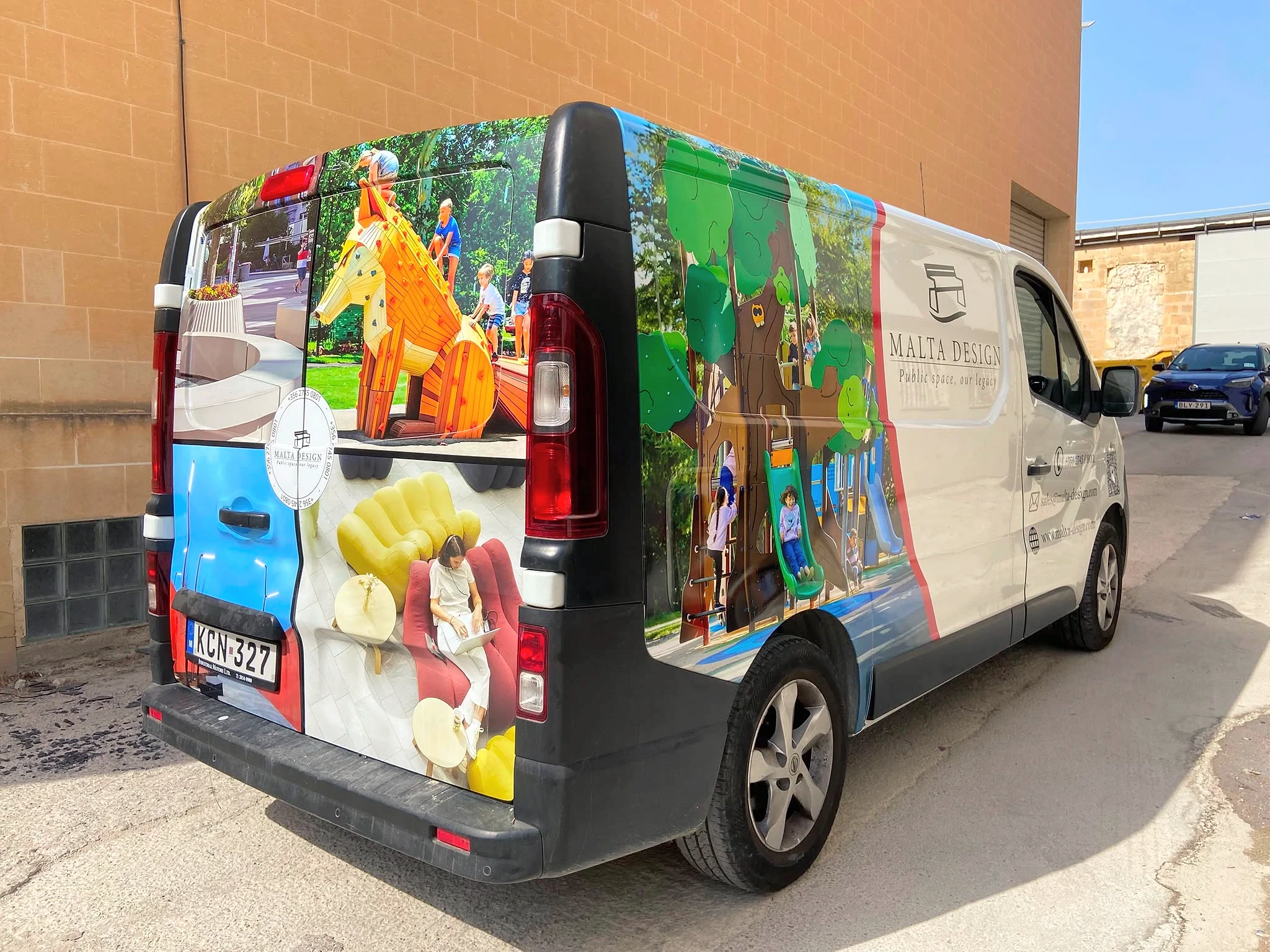 White panel van wrapped in photographs of outdoor play equipment and interior furniture, parked beside a stone wall