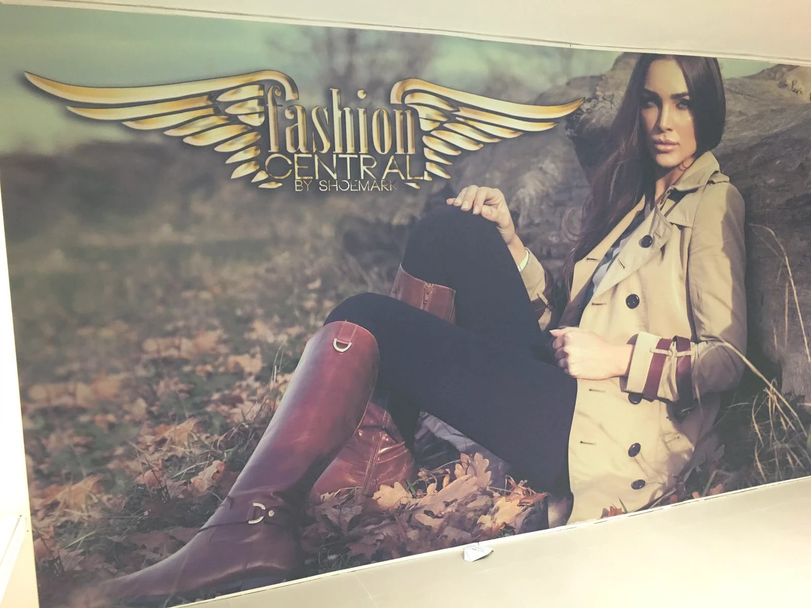 Wall-sized photographic print of a model in a trench coat and riding boots seated among autumn leaves, with a gold winged emblem above