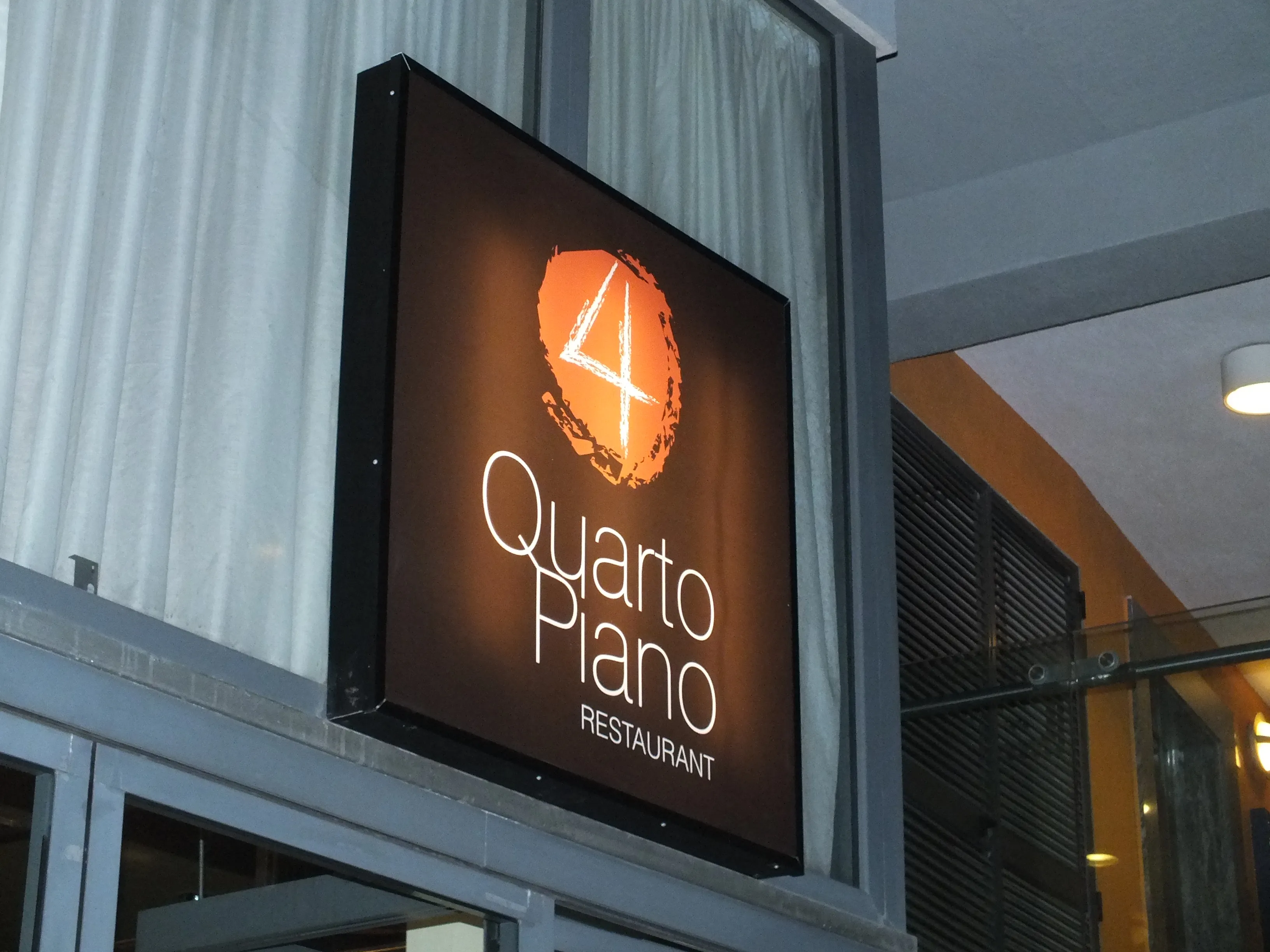 Internally lit blade sign in a dark tray frame, mounted on brackets perpendicular to a building wall above an entrance