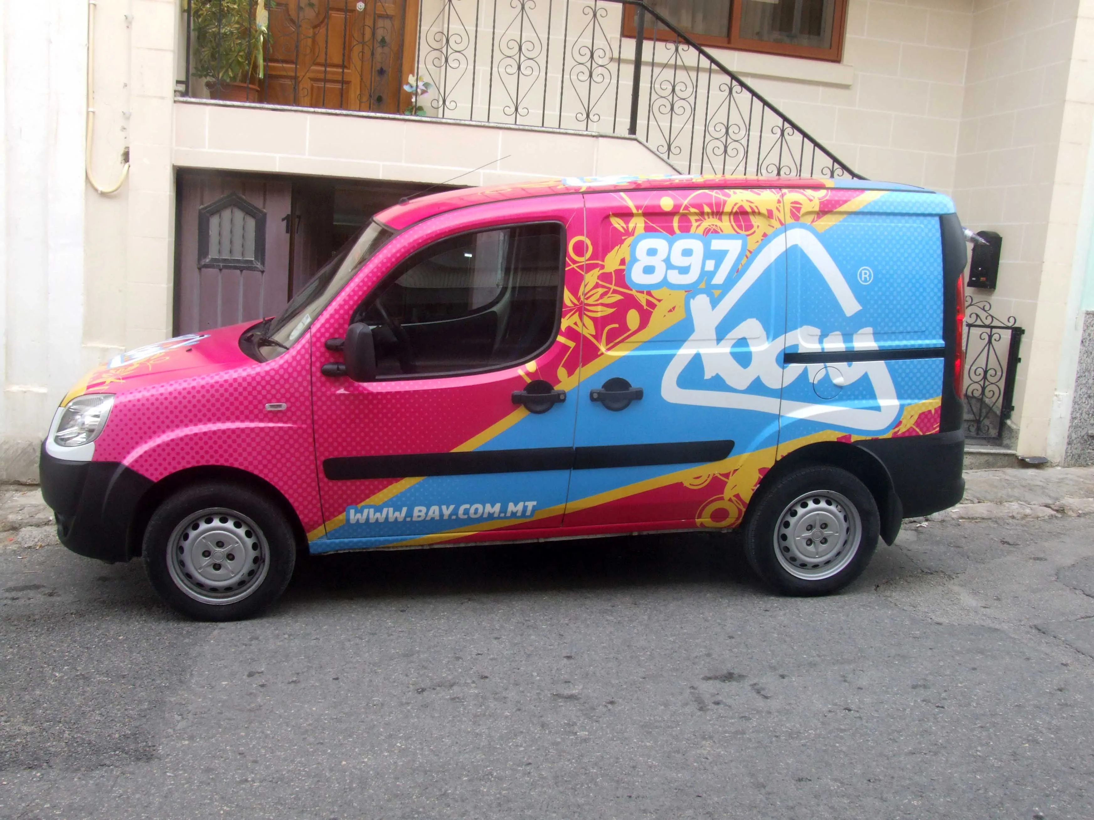 Compact van wrapped magenta at the front through blue at the rear, with a diagonal flash, patterned ground, a large station mark and a web address along the sill