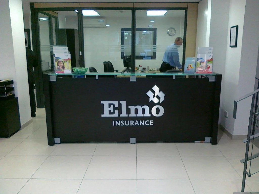 Dark-clad reception counter with a glass transaction shelf on brackets, carrying a dimensional metal logo mark and company name on its front face