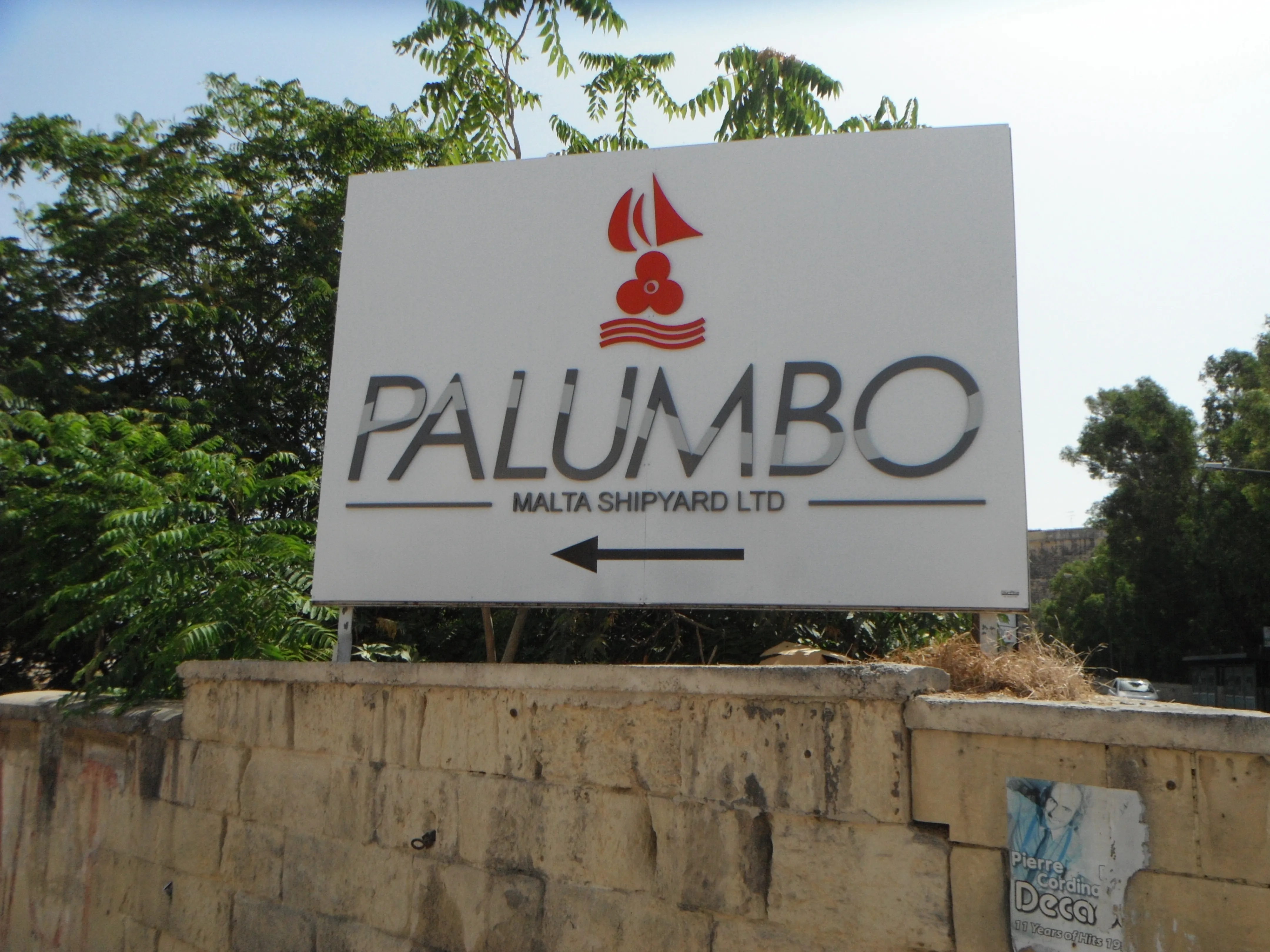 Large white directional sign on posts above a stone boundary wall, carrying a red logo, dimensional metal letters, a descriptor line and an arrow
