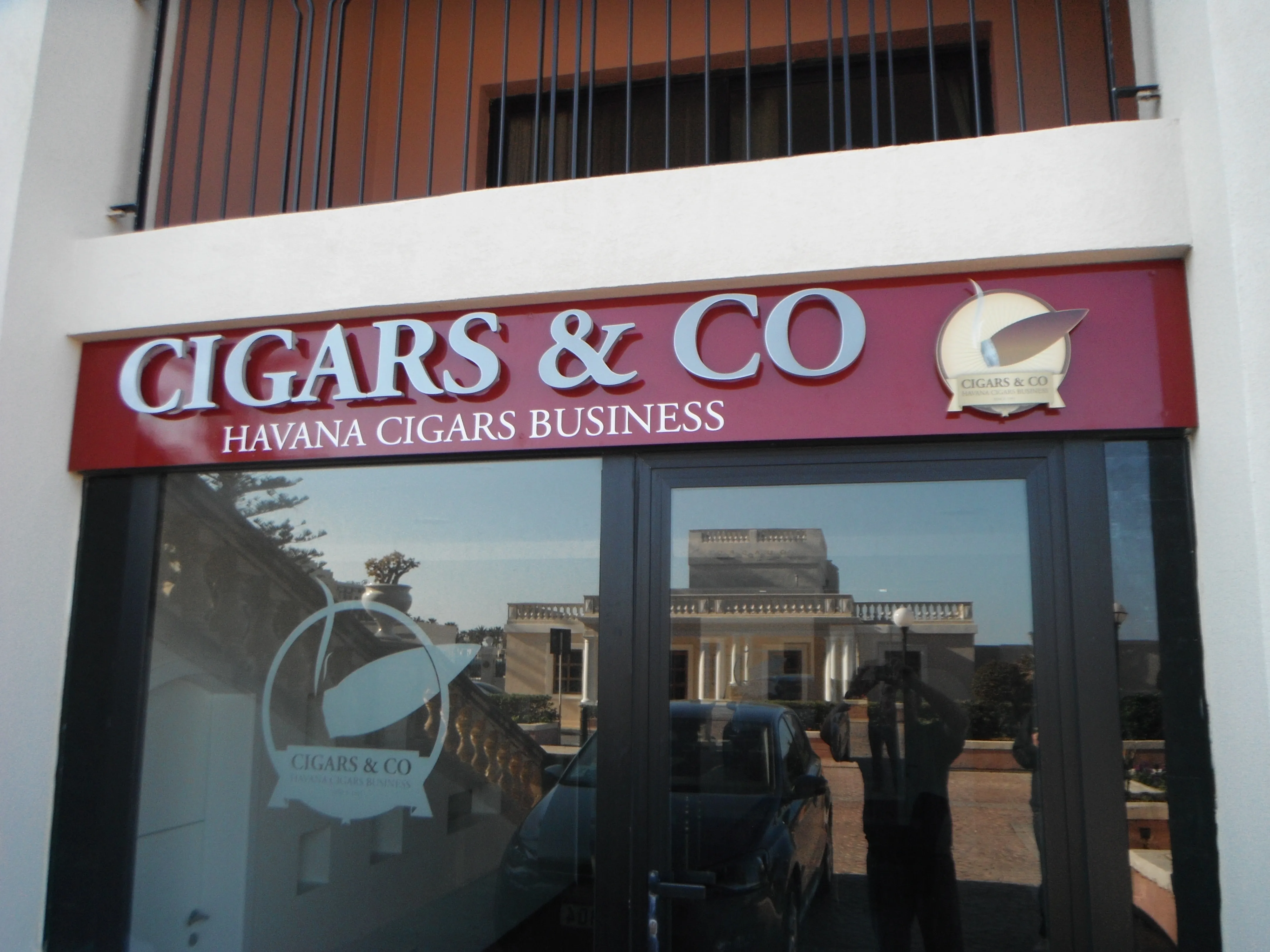 Deep red shopfront fascia carrying brushed metal dimensional letters and a circular badge, above glazing bearing an etched version of the same badge