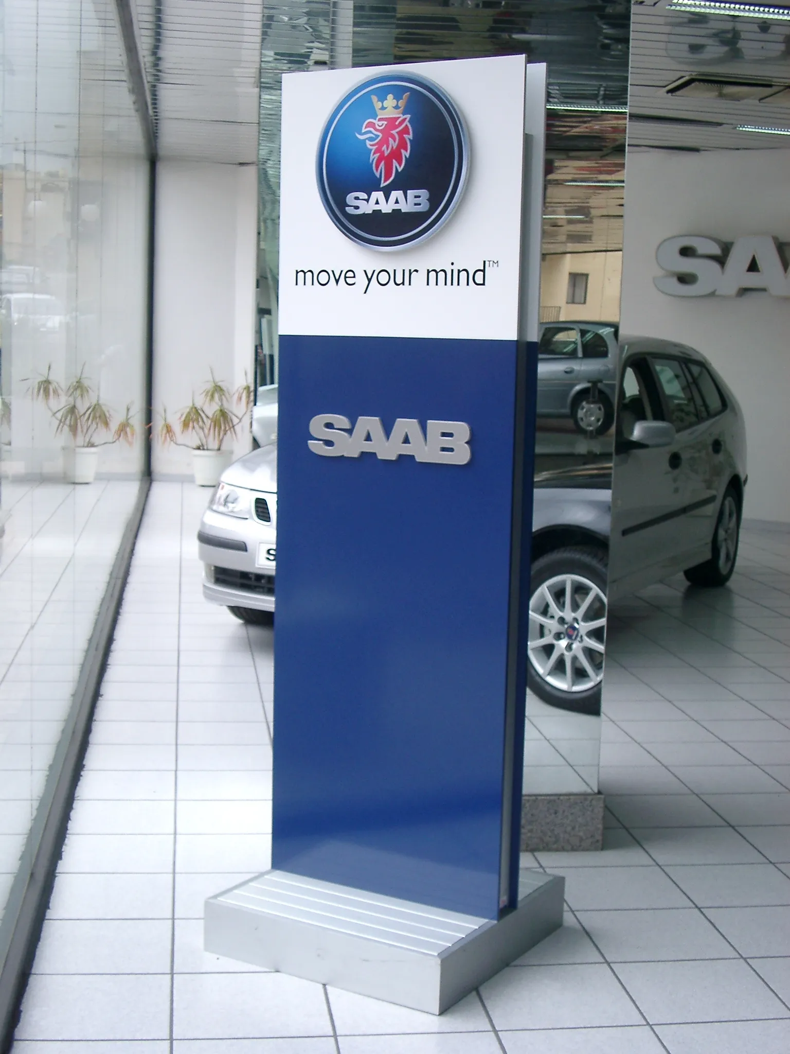 Free-standing two-tone totem sign on a stepped brushed-metal base in a car showroom, carrying a dimensional badge, a strapline and metal-faced letters