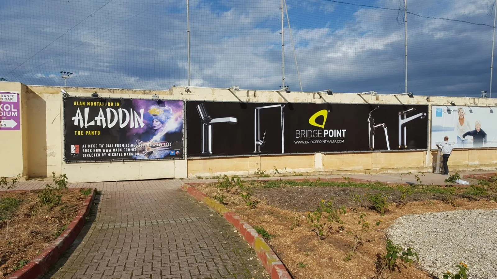 Printed banner fitted along a site hoarding beside a road, with rough ground in front