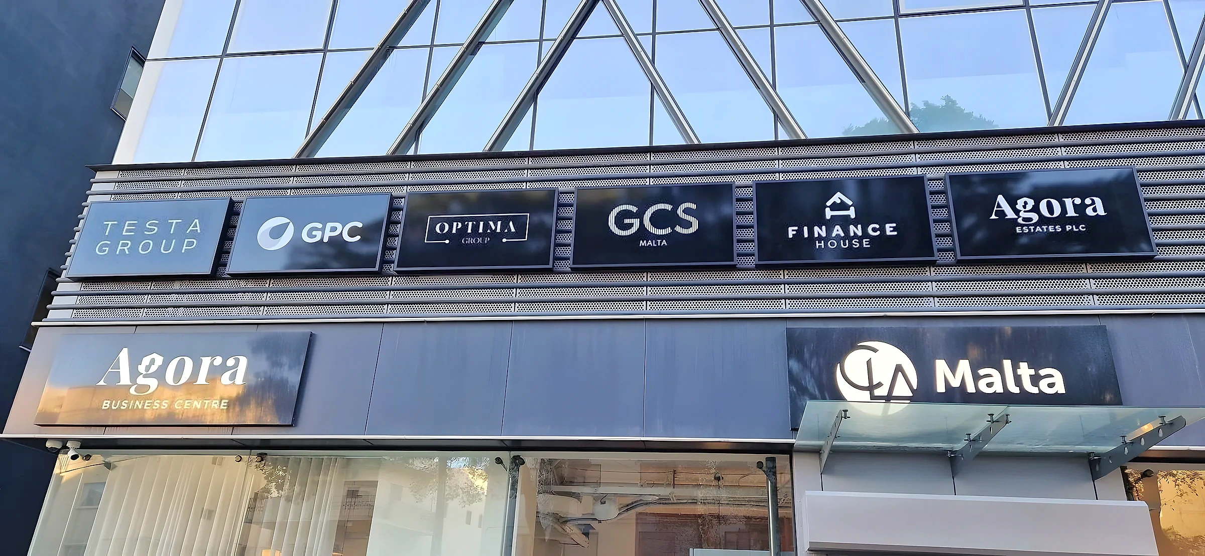 Row of six dark tenant name panels above two larger illuminated signs on a glazed building frontage