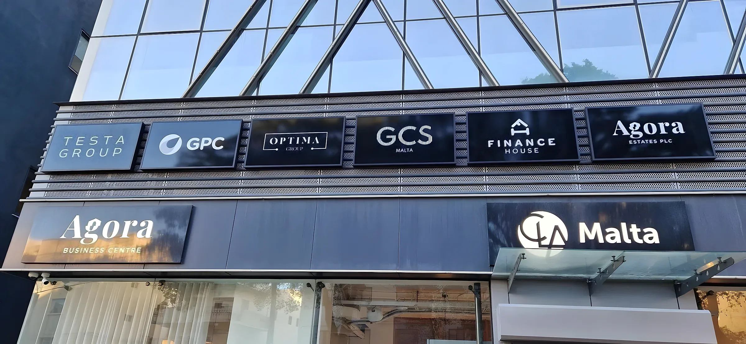Row of six dark tenant name panels above two larger illuminated signs on a glazed building frontage