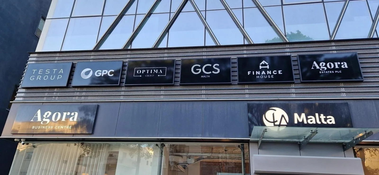 Row of six dark tenant name panels above two larger illuminated signs on a glazed building frontage